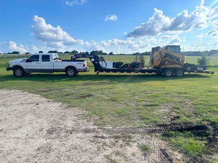 White pickup truck towing a yellow skid steer on a flatbed trailer across a grassy field under a blue sky