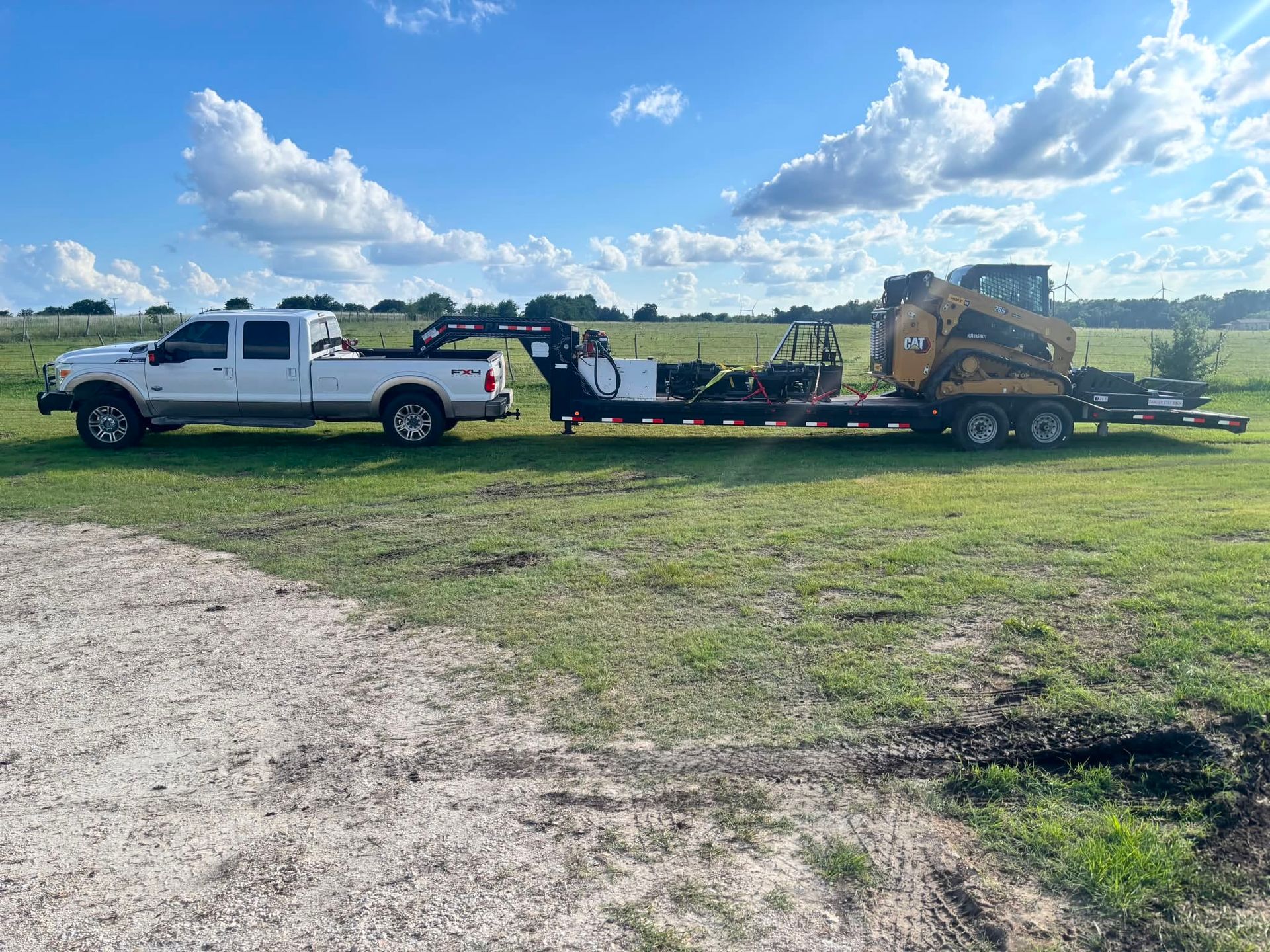 White pickup truck towing a flatbed trailer with equipment across a grassy field under a blue sky