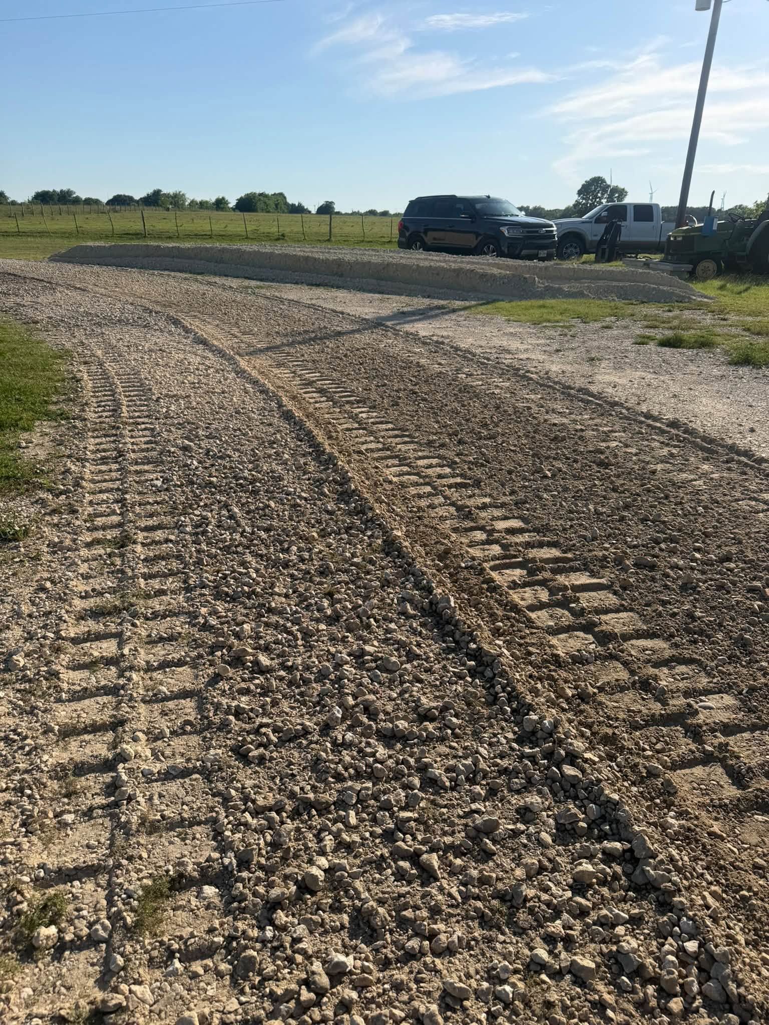 Gravel farm road with tire tracks leading to a pickup truck in a sunny field under a clear sky