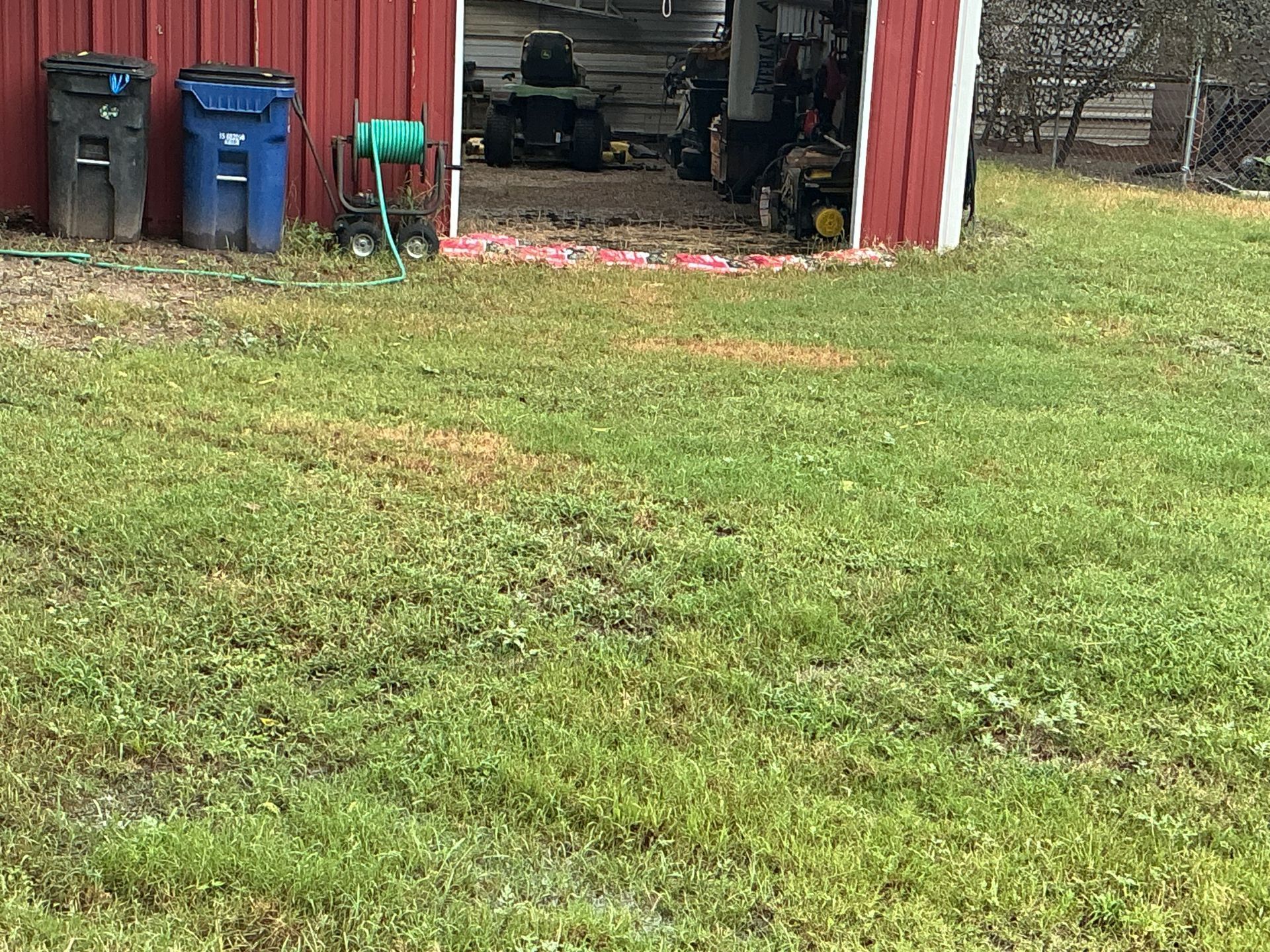 Grassy yard beside a red shed with trash bins, a green chair, and a partially open garage.