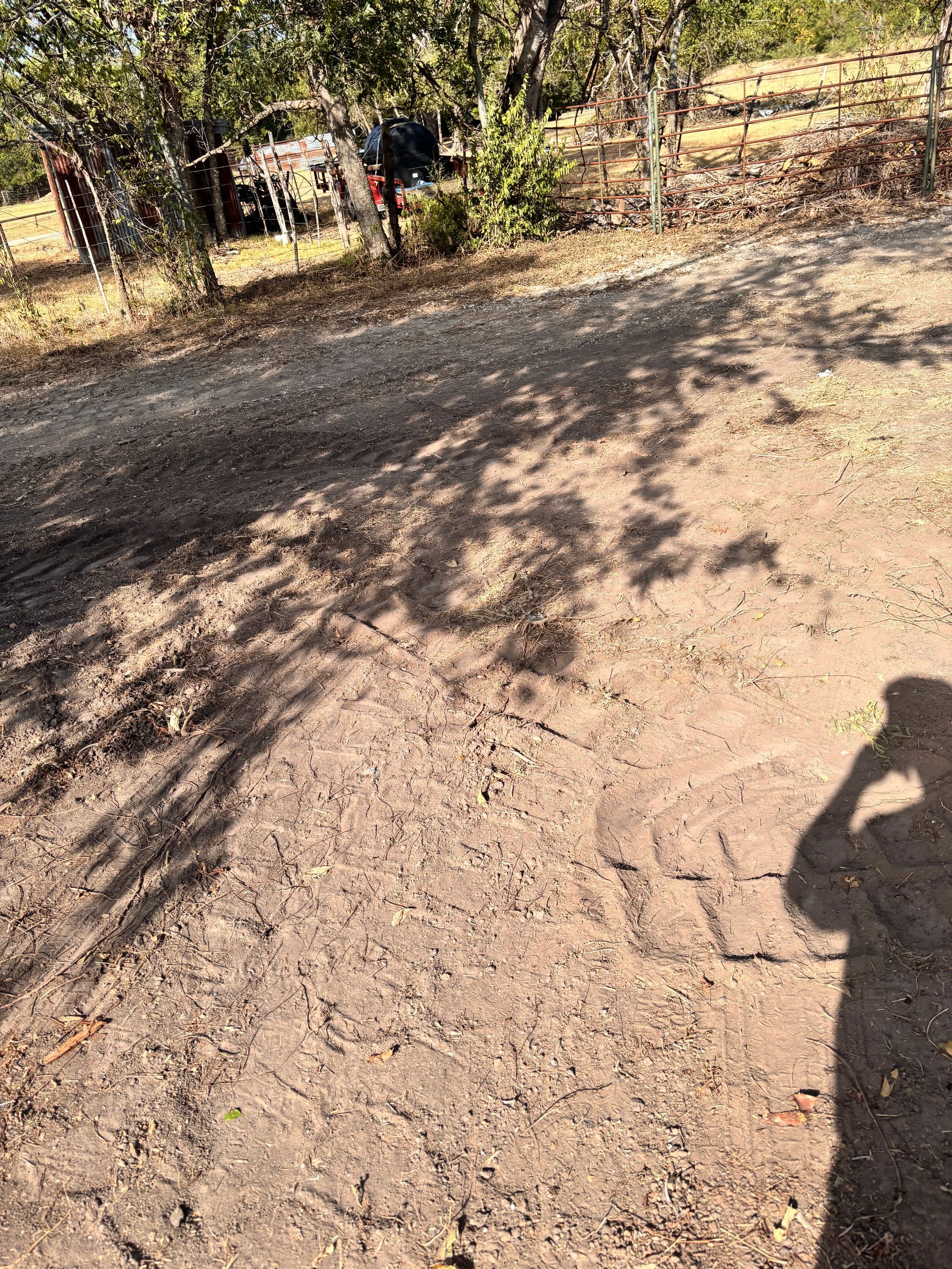 Dusty dirt path with trees and shadows; a person stands near a gate in the background.