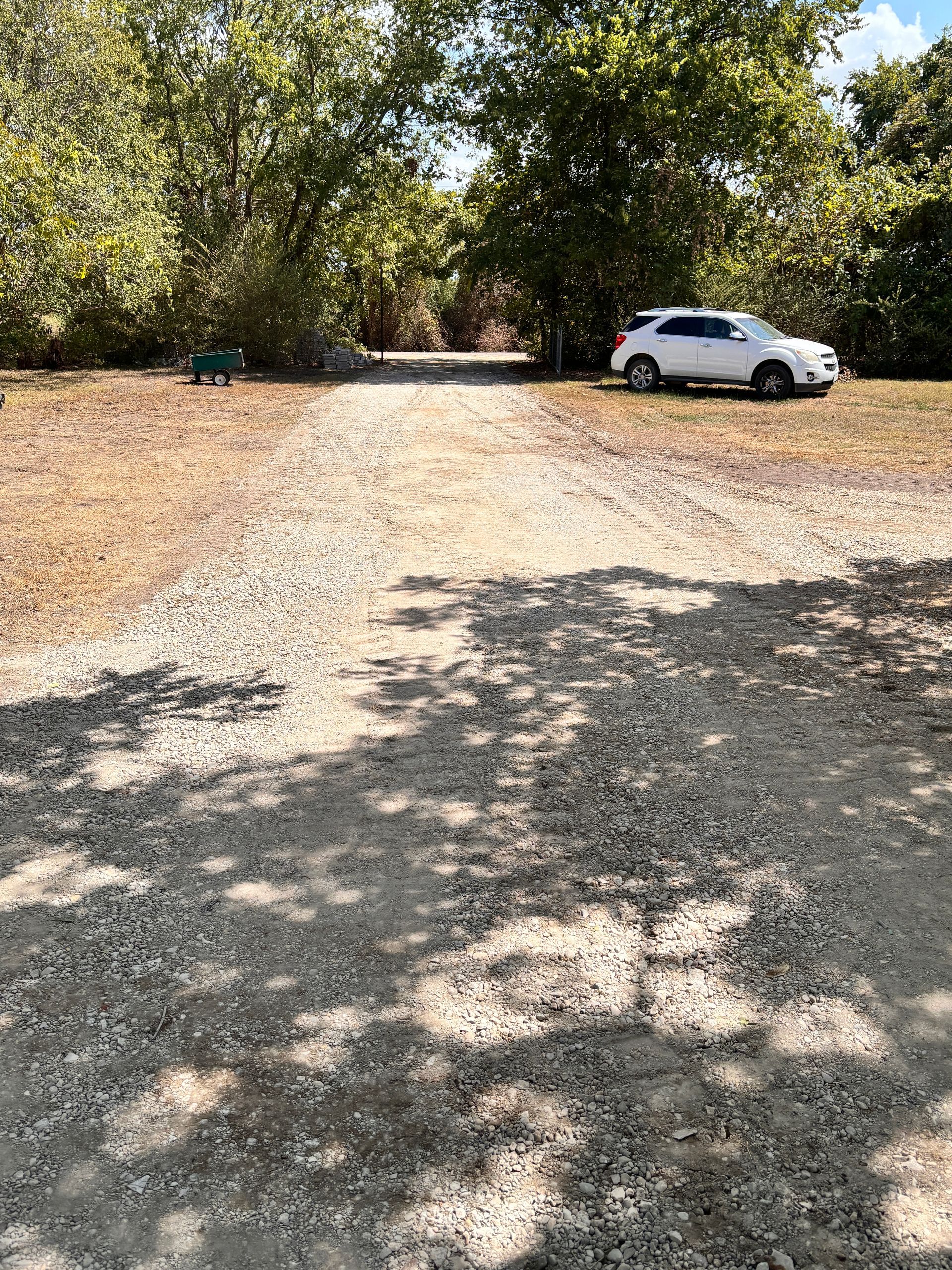 Gravel driveway under trees with a white car parked on the right in bright sunlight.