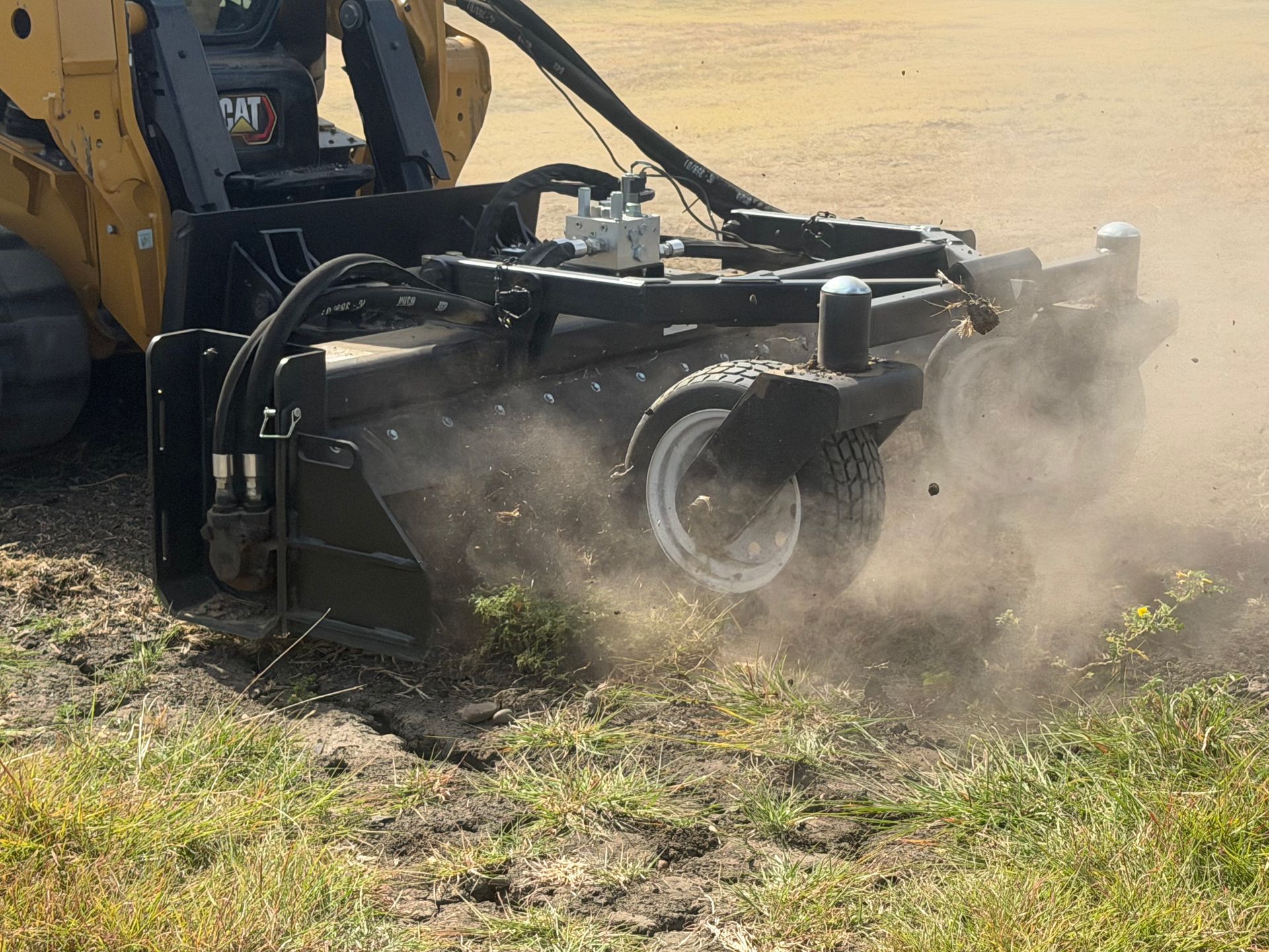 Yellow excavator using a brush cutter attachment to clear dusty ground near grass.