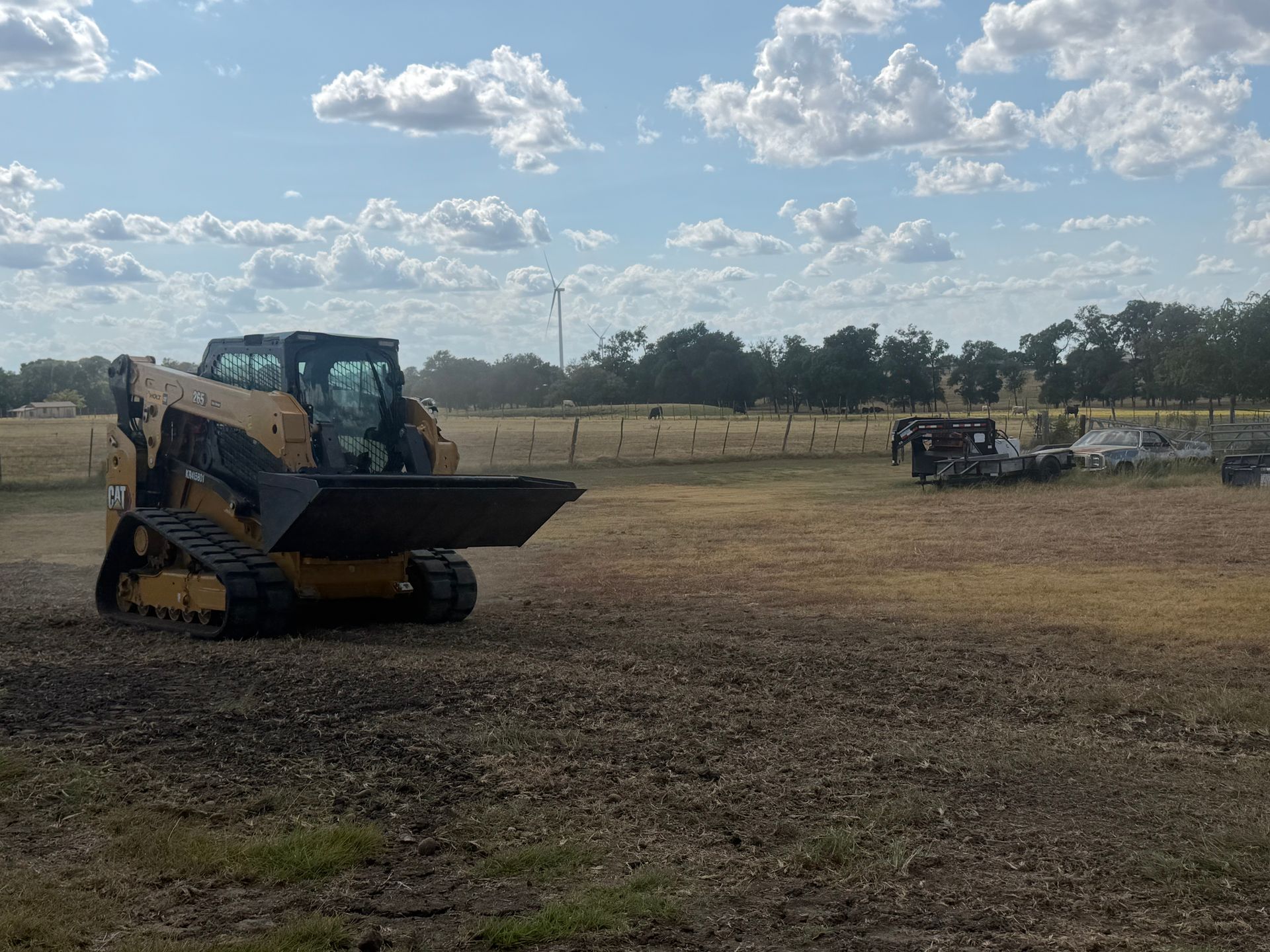Yellow skid steer loader on a dirt field under a cloudy sky, with fencing and equipment in the background