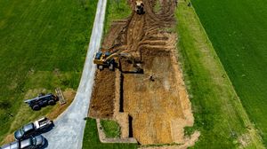 Aerial view of construction equipment grading a dirt strip beside a green field and road