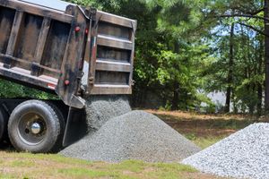 Dump truck unloading gray gravel onto a pile beside a wooded roadside.