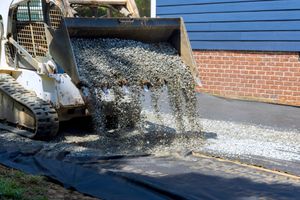 Construction machine pouring gravel onto a road beside a brick wall