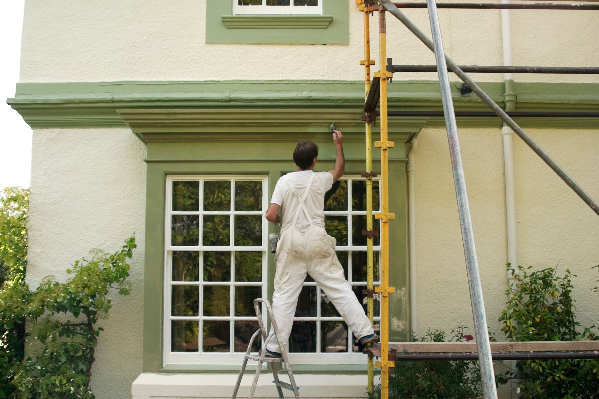 Painter on a ladder, painting green trim on a white building exterior.
