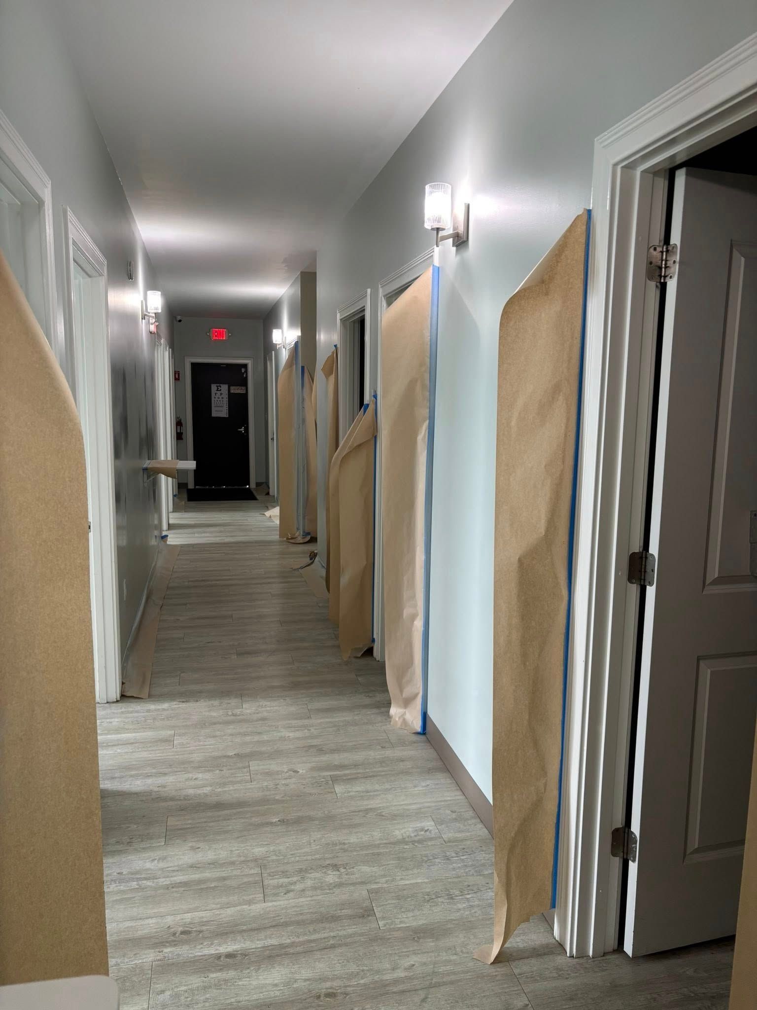 Long hallway with closed doors, some covered in brown paper, gray flooring, and overhead lighting.