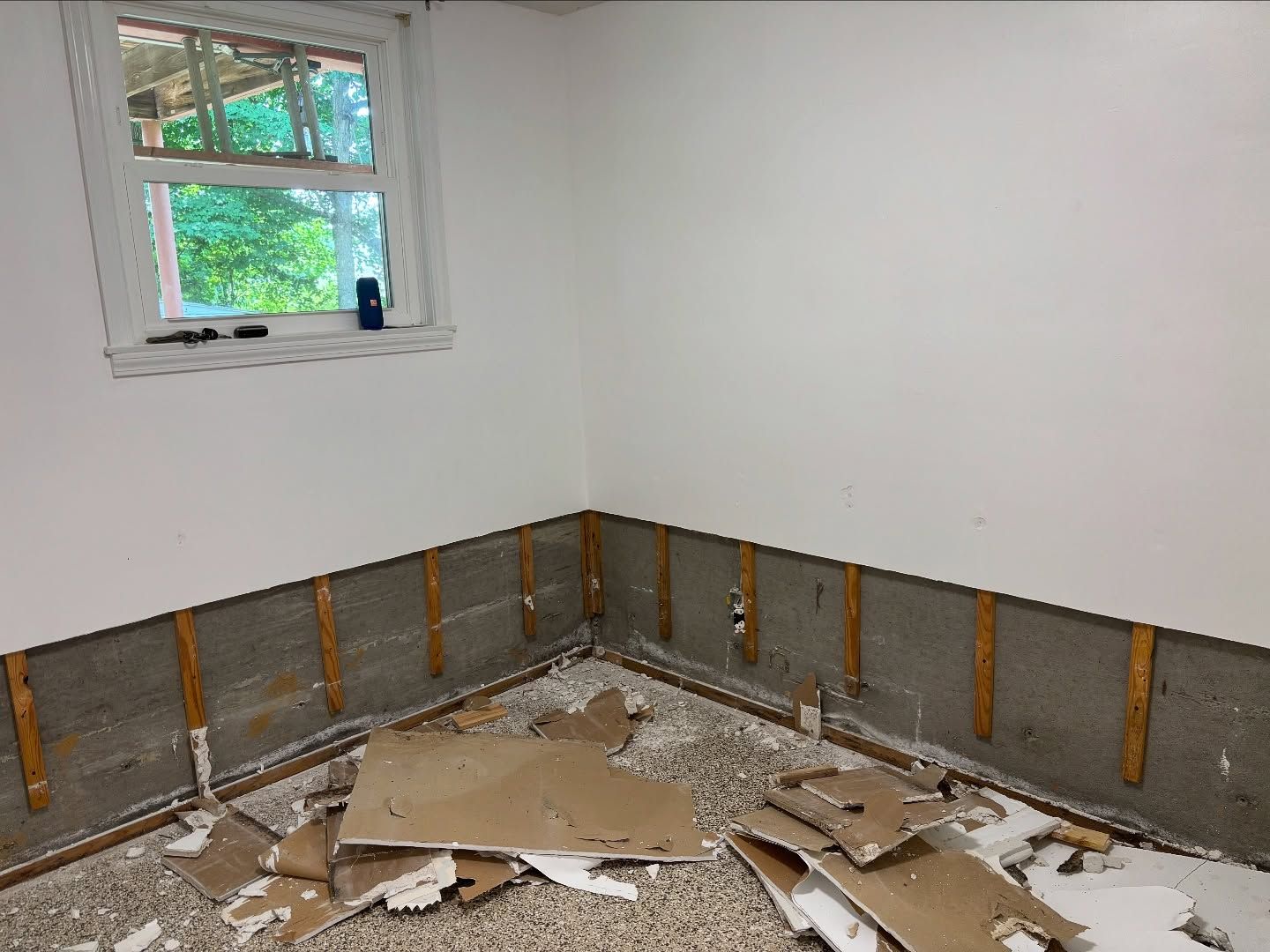 Room with exposed concrete and wooden strips along the lower walls, window in the corner, debris on the floor.
