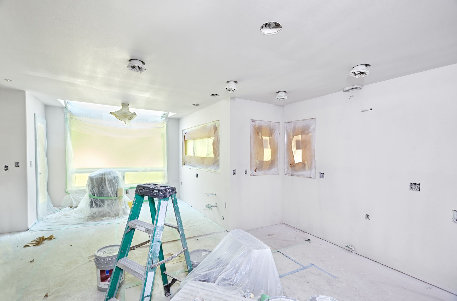 Room being renovated, with white walls, ladder, covered furniture, and masked windows.