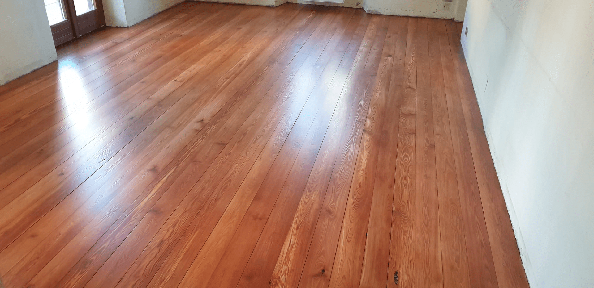 Pavimento in legno lucidato con lunghe assi che riflettono la luce in una stanza dalle pareti bianche.