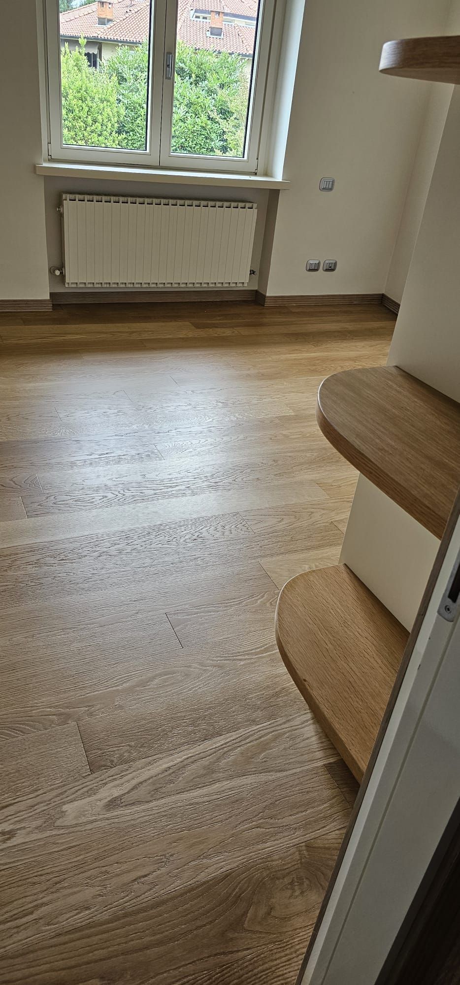 Pavimenti in legno massello in una stanza con finestra, termosifone e scale.