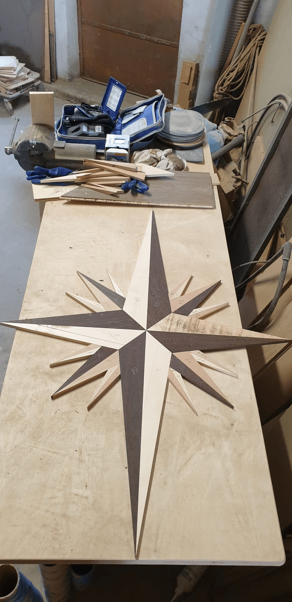 Intarsio a forma di rosa dei venti in legno chiaro e scuro su una superficie di compensato, in un ambiente di laboratorio.