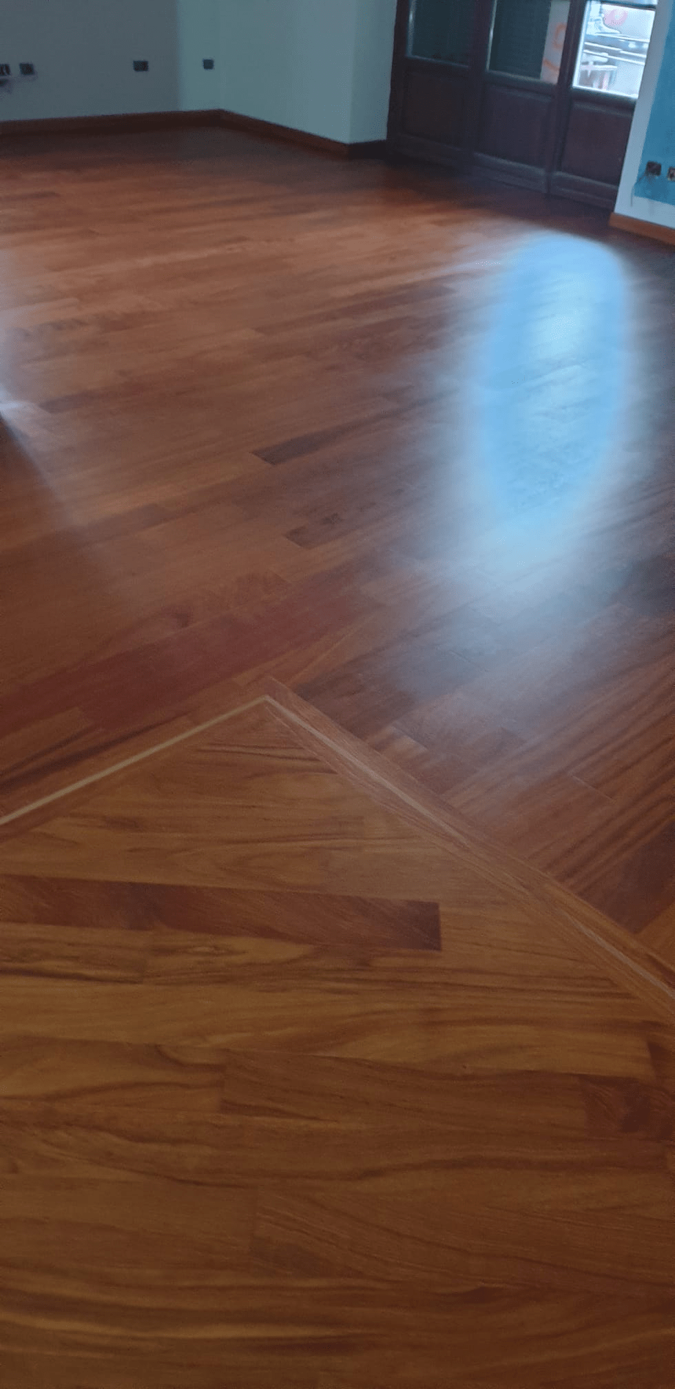 Pavimento in legno con superficie riflettente. Bordo triangolare visibile. Stanza con porta e finestra.