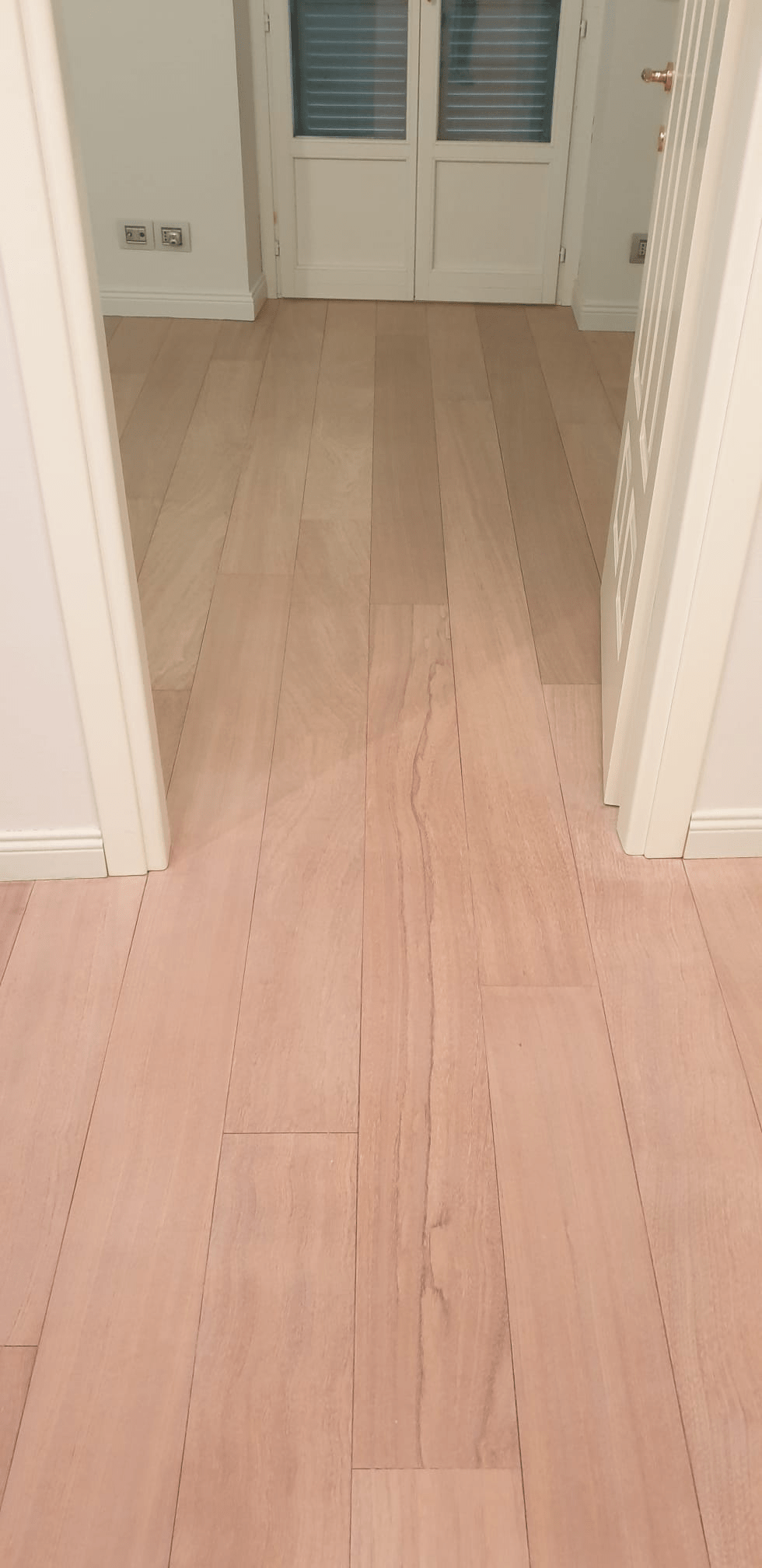 Pavimento in legno chiaro in un corridoio che conduce a una porta bianca con finestra. Le pareti bianche incorniciano lo spazio.