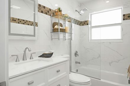 A bright white bathroom with a vanity, marble-patterned tiled walls, a decorative mosaic border, and a glass-enclosed tub.