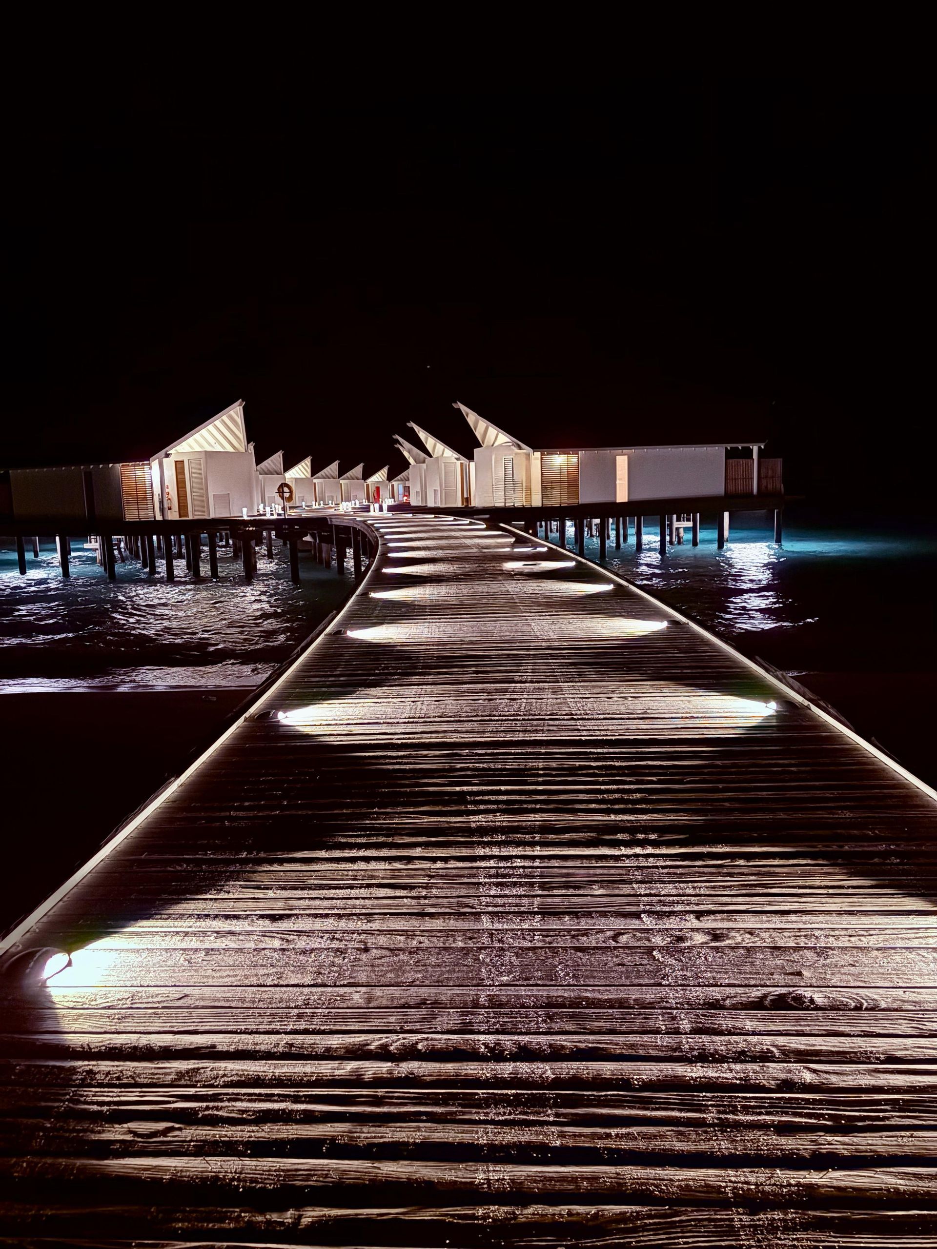 Wooden pier with lights leading to lit huts over dark water at night.