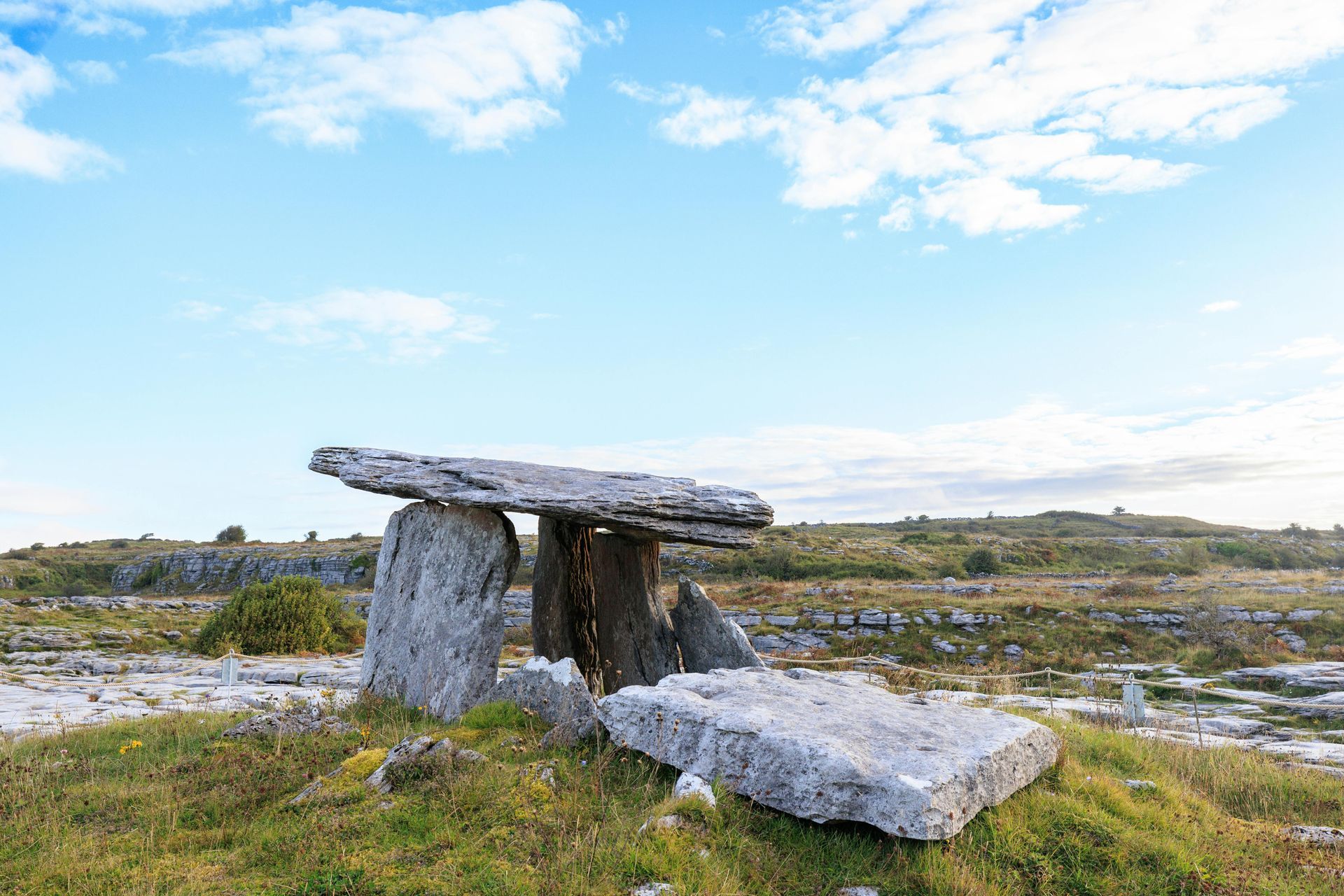 Dolmen structure made of large stones under a blue sky, in a grassy field.
