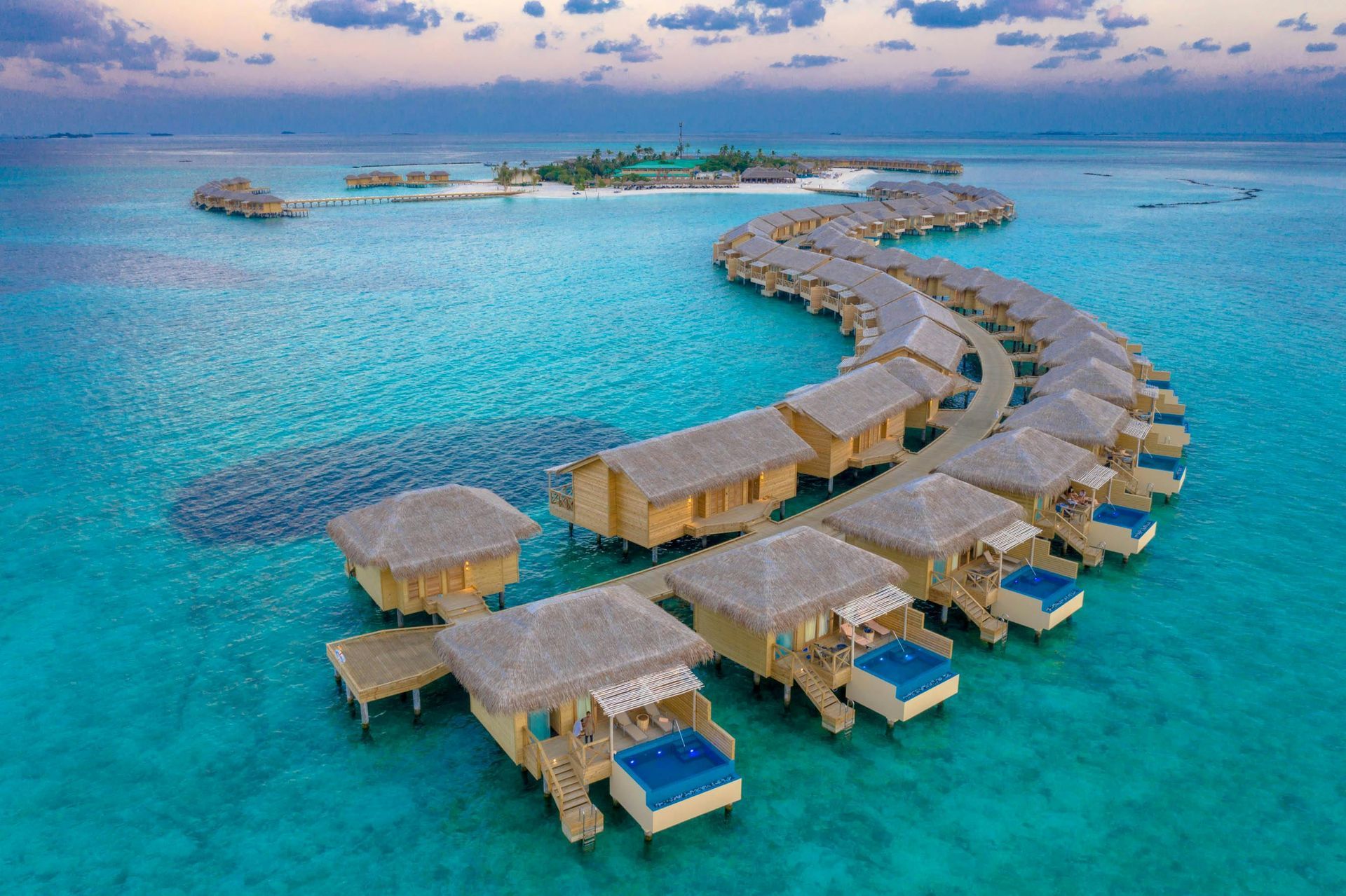 Overwater bungalows in a turquoise ocean, forming a curved line to a green island in the distance.