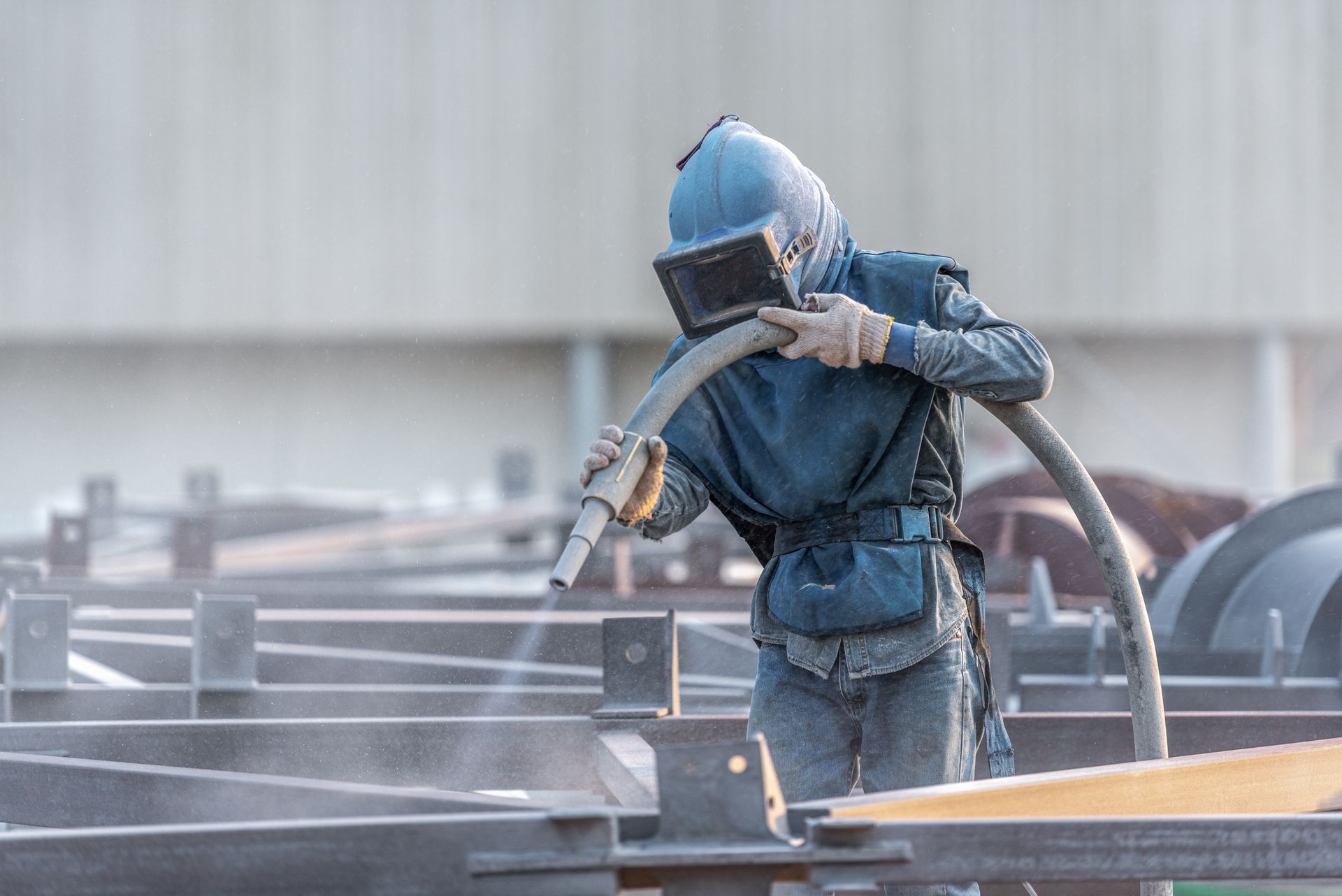 Worker in protective gear sandblasting metal surface with high pressure hose.