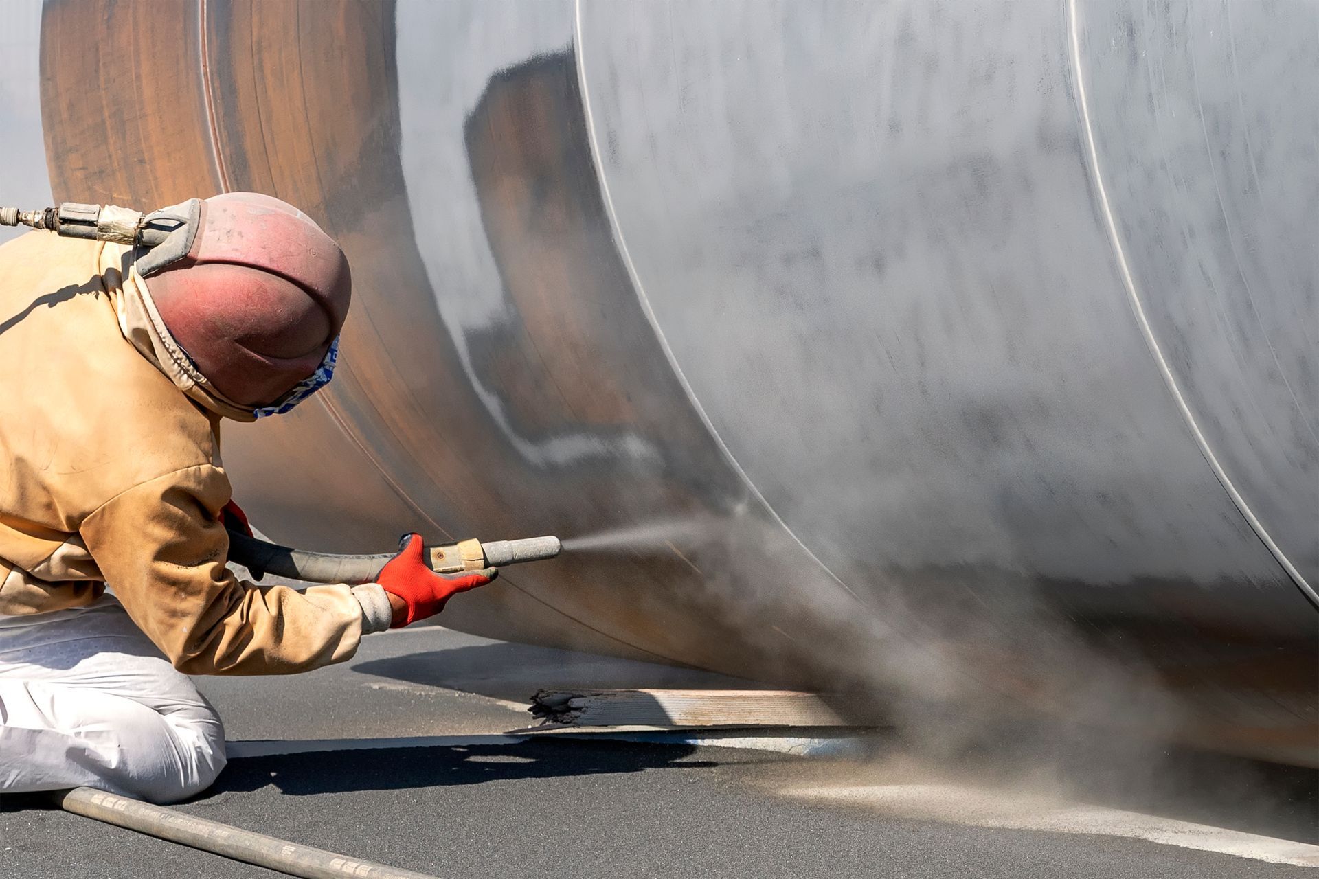 Worker in protective gear sandblasting a large metal tank with a high-pressure hose. Worker in protective gear sandblasting a large metal tank with a high-pressure hose.