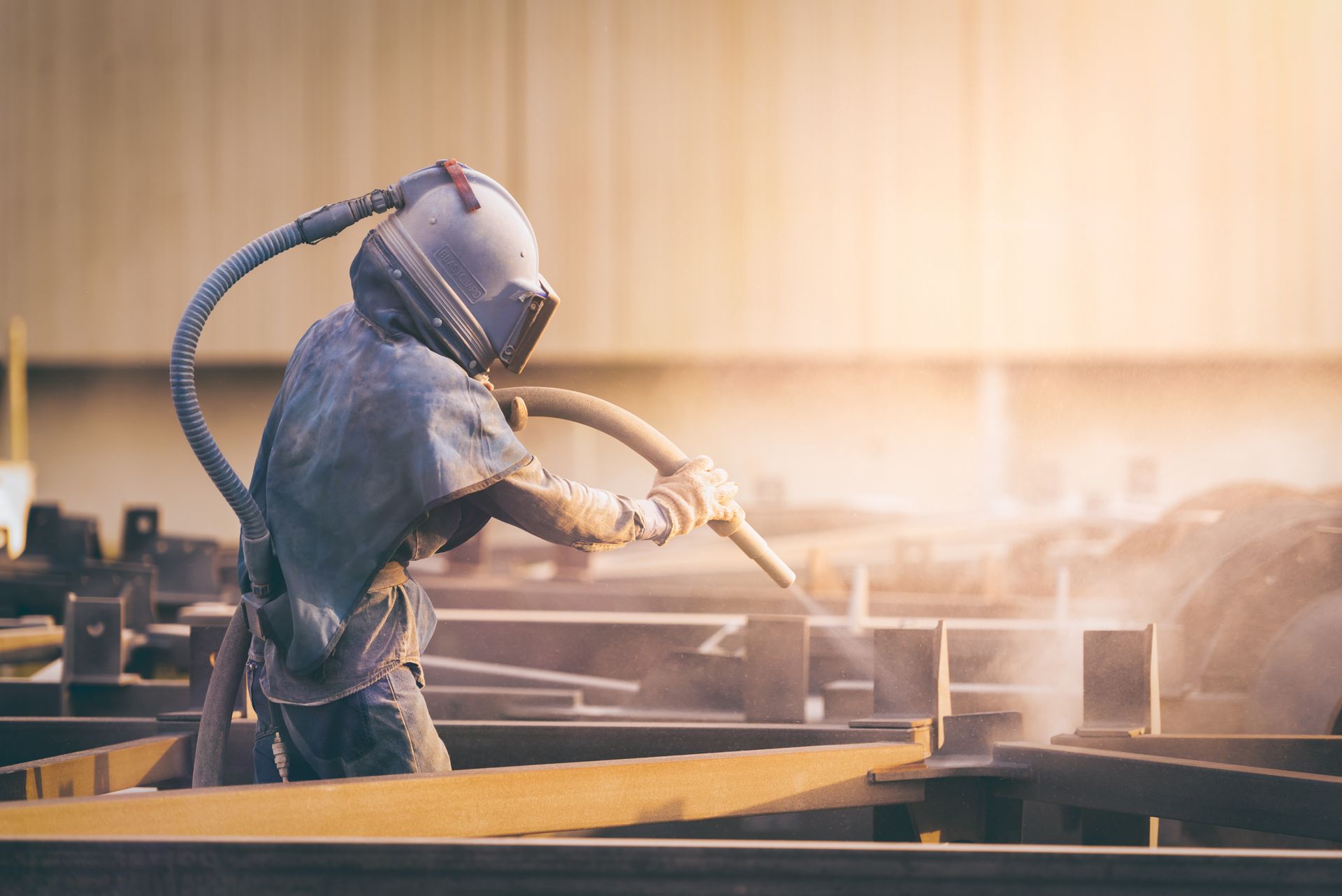 Worker in protective hood abrasive blasting steel beams in a workshop. Worker in protective hood abrasive blasting steel beams in a workshop.
