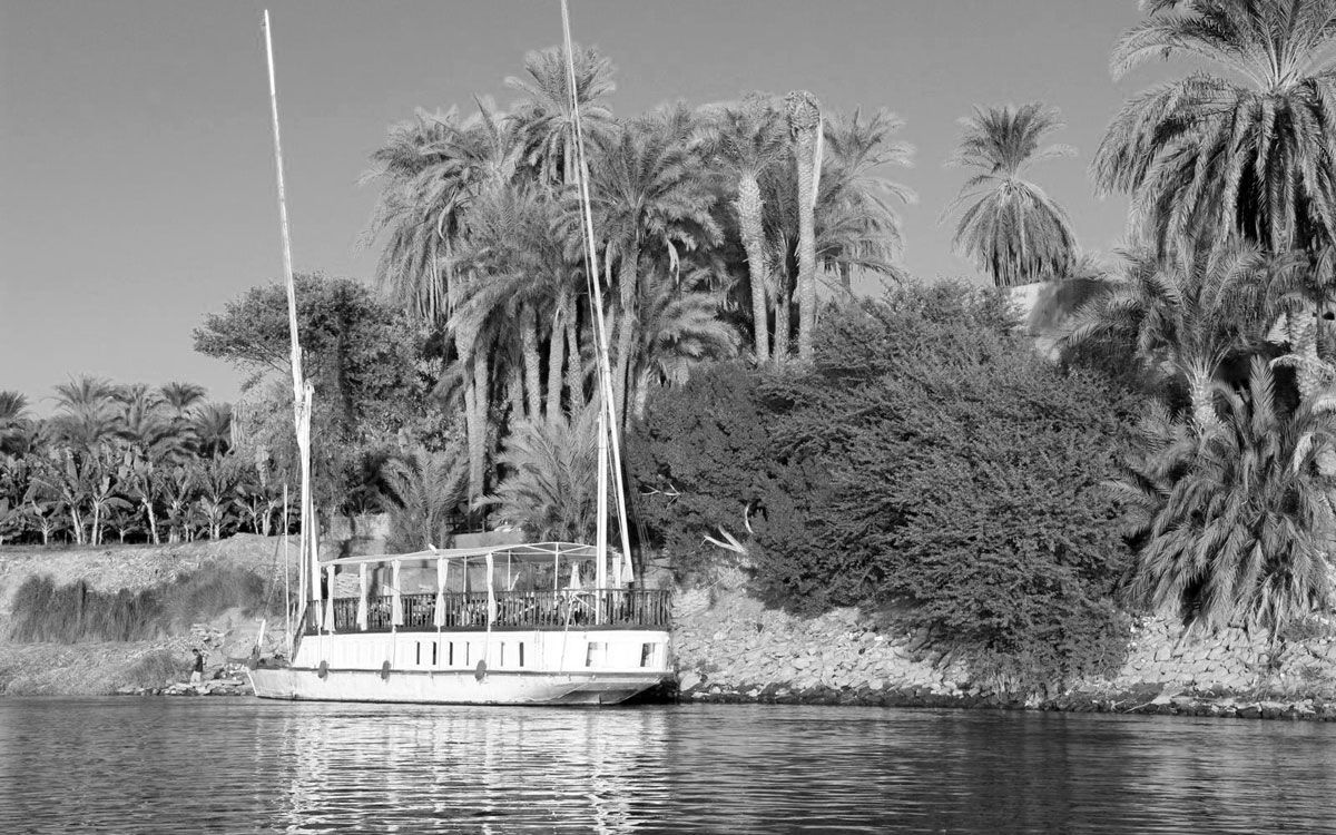 A historical photo of a Dahabiya Kingfisher sailing on the Nile in Egypt in 1899. The photo is in black and white and shows the boat from a side view. The Dahabiya Kingfisher is a traditional wooden boat that can be rented for private cruises between Luxor and Aswan.