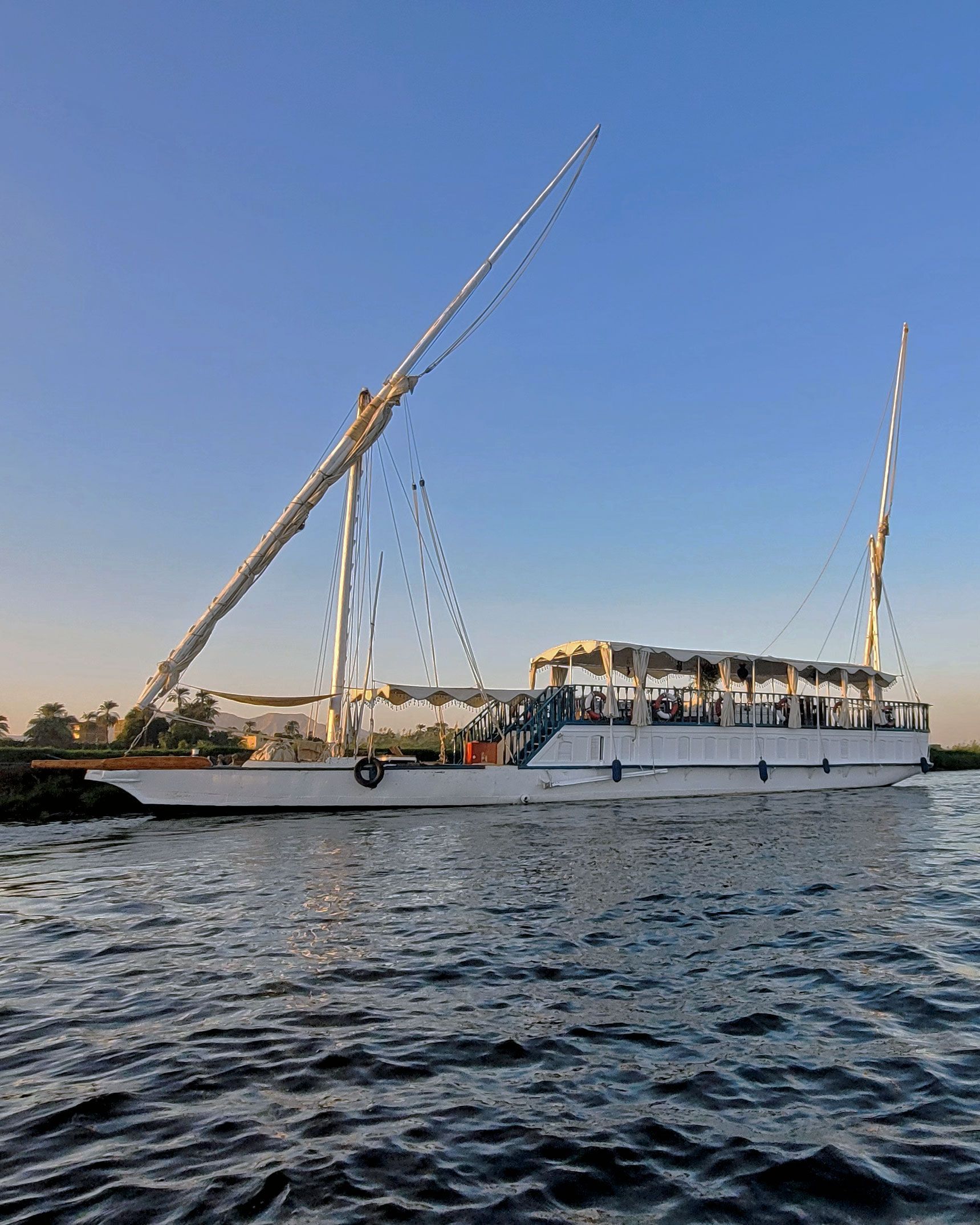 In this photo, you can see the Dahabiya Kingfisher, a traditional wooden boat that dates back to 1899. The boat has a graceful white hull, it sails along the Nile river, the longest and most ancient river in the world. The boat offers private cruises for travelers who want to experience the beauty and culture of the Nile valley, where they can visit temples, tombs, and villages along the way.