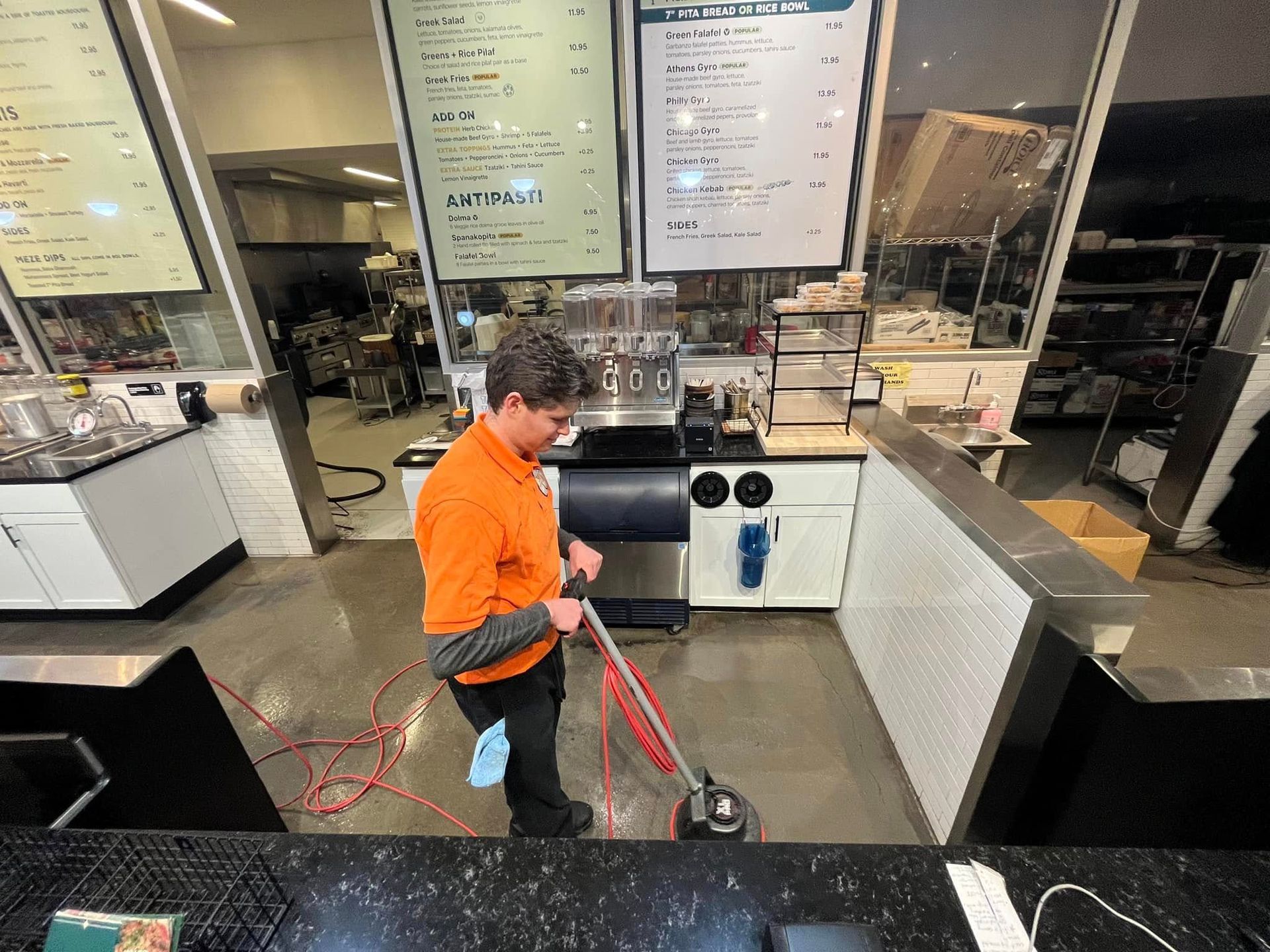 Worker in orange shirt cleaning a fast-food restaurant floor with a mop near the counter