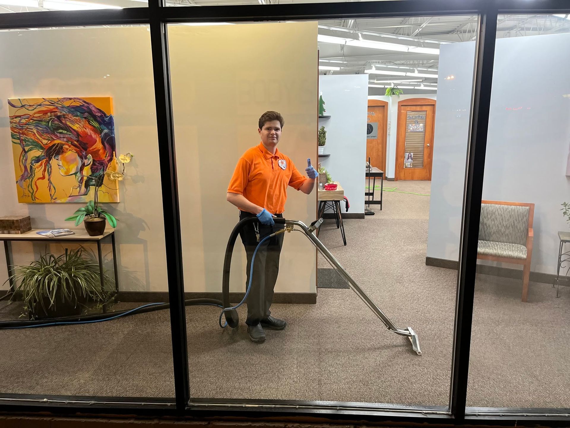 Worker vacuuming carpet in office hallway through a glass window