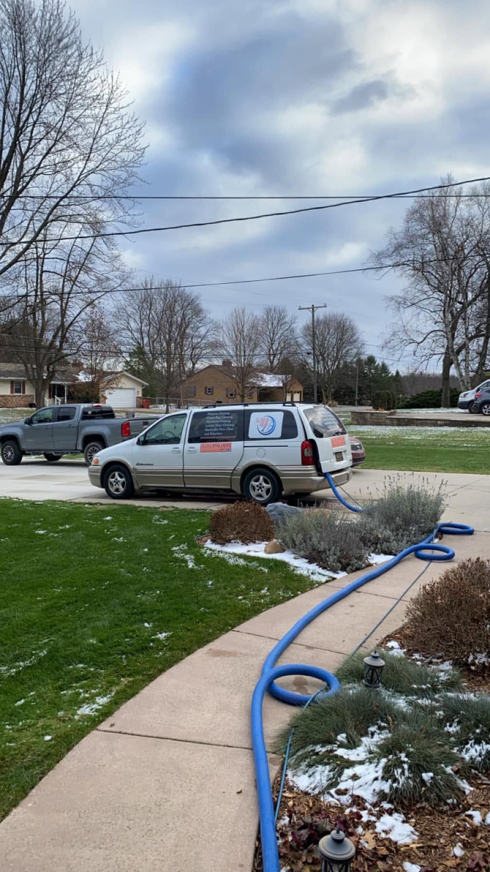 Utility van parked on a suburban lawn beside a sidewalk, with a blue hose and patches of snow nearby.