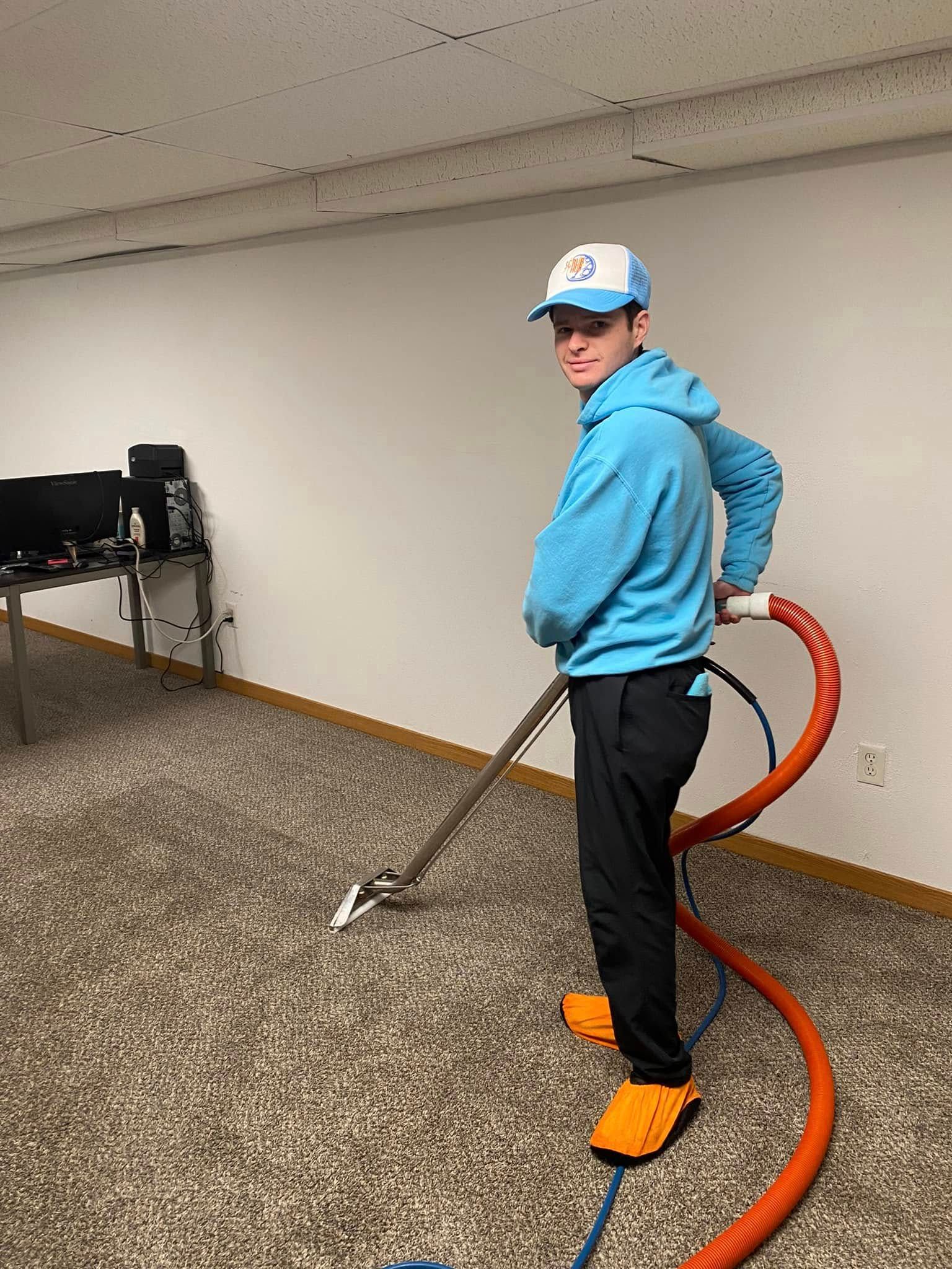 Person using a floor-cleaning machine in a carpeted room, wearing a blue hoodie and orange shoe covers.