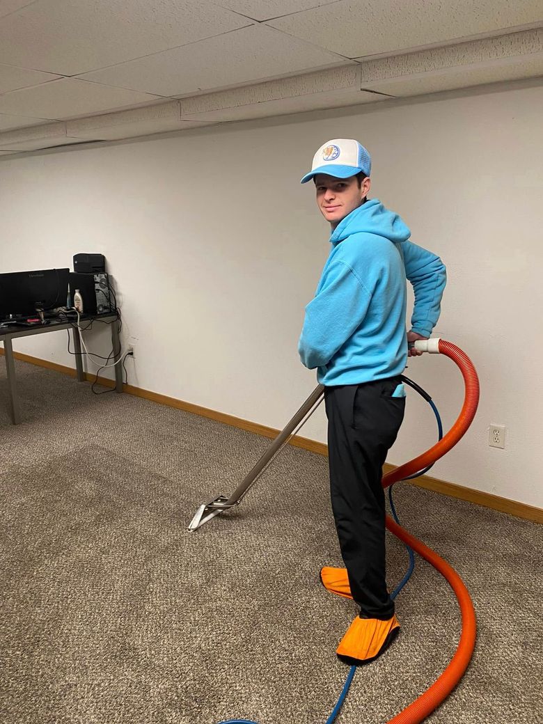 Person using a floor-cleaning machine in a carpeted room, wearing a blue hoodie and orange shoe covers.