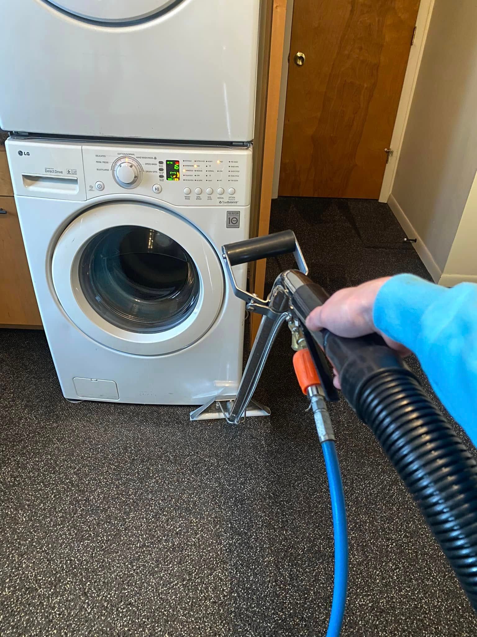 Hand vacuum hose cleaning the front door of a white washing machine in a laundry room