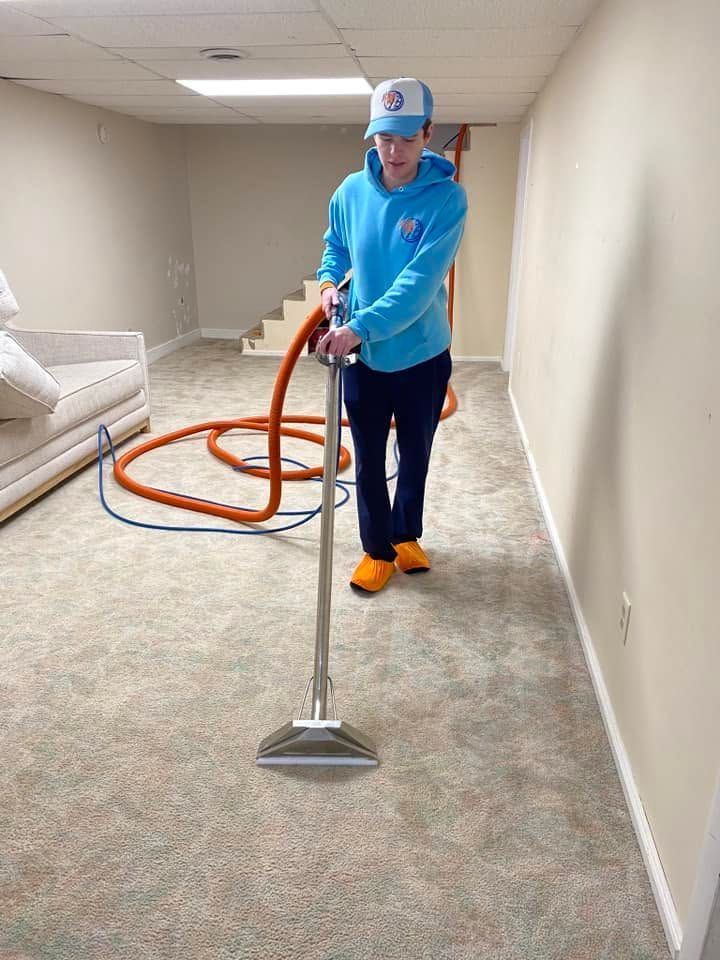 Person vacuuming beige carpet in a basement, wearing blue shirt and orange shoes.