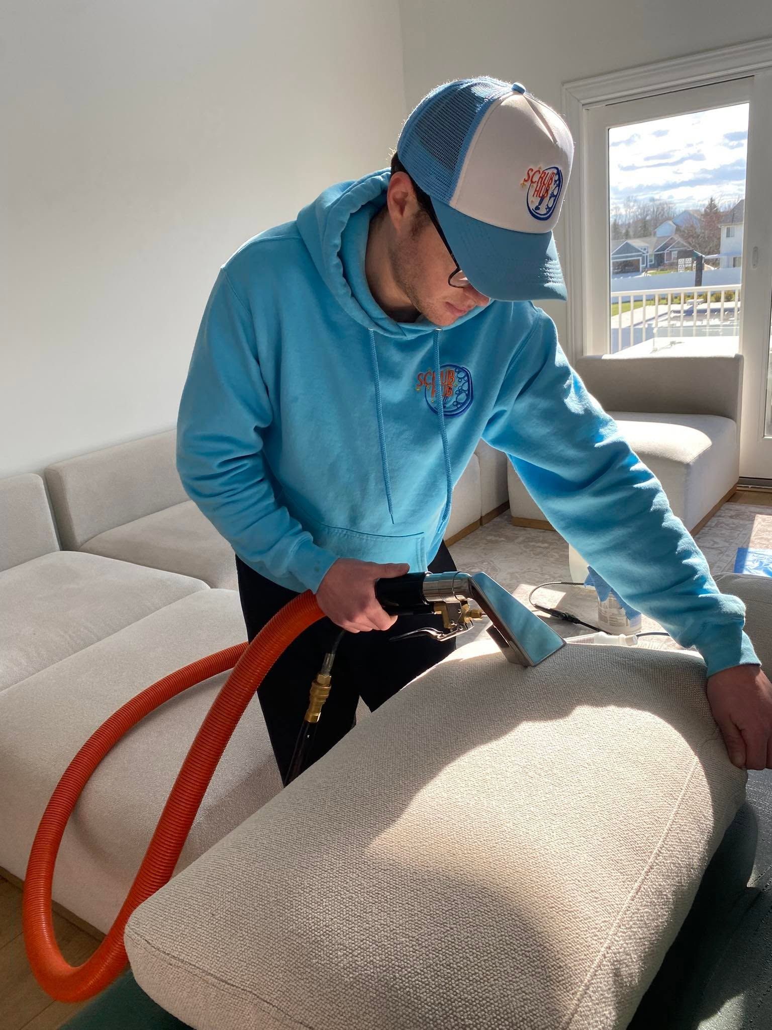 Person using a vacuum cleaner on a beige sofa in a bright living room
