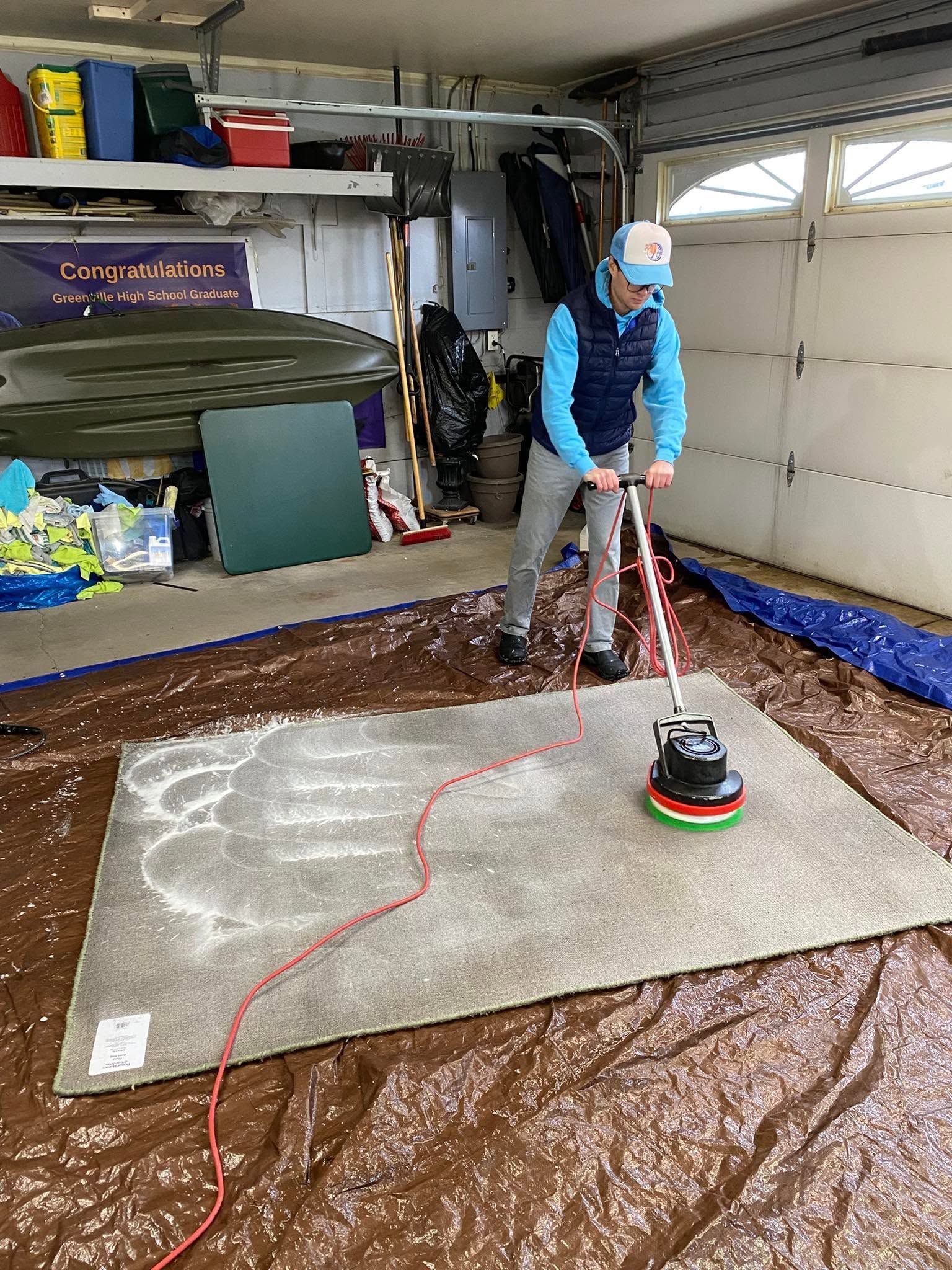 Person polishing a large carpeted rug with a floor buffer in a garage workshop