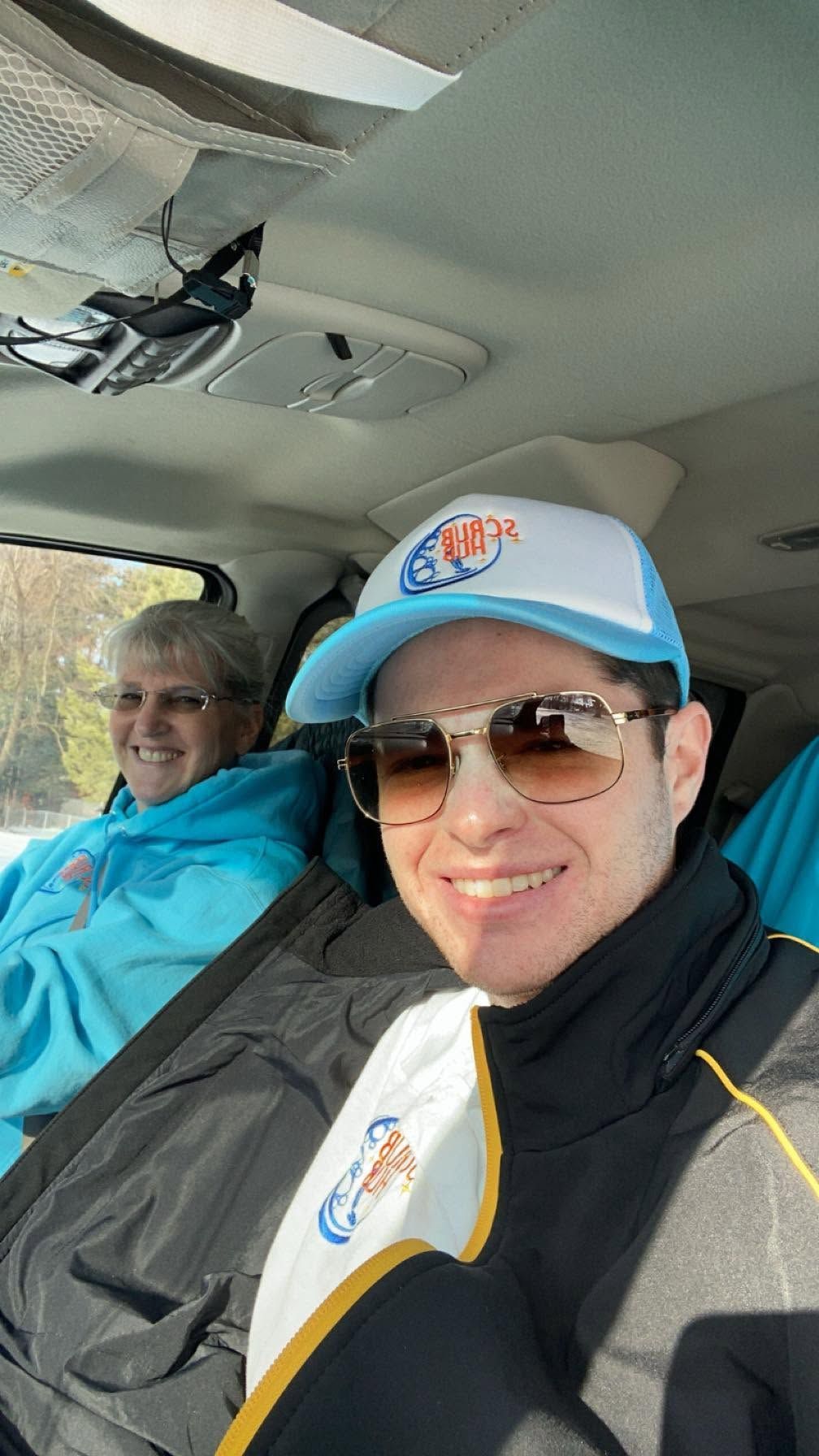 Two people in a car, smiling in jackets and caps, with a partially open sunroof above.