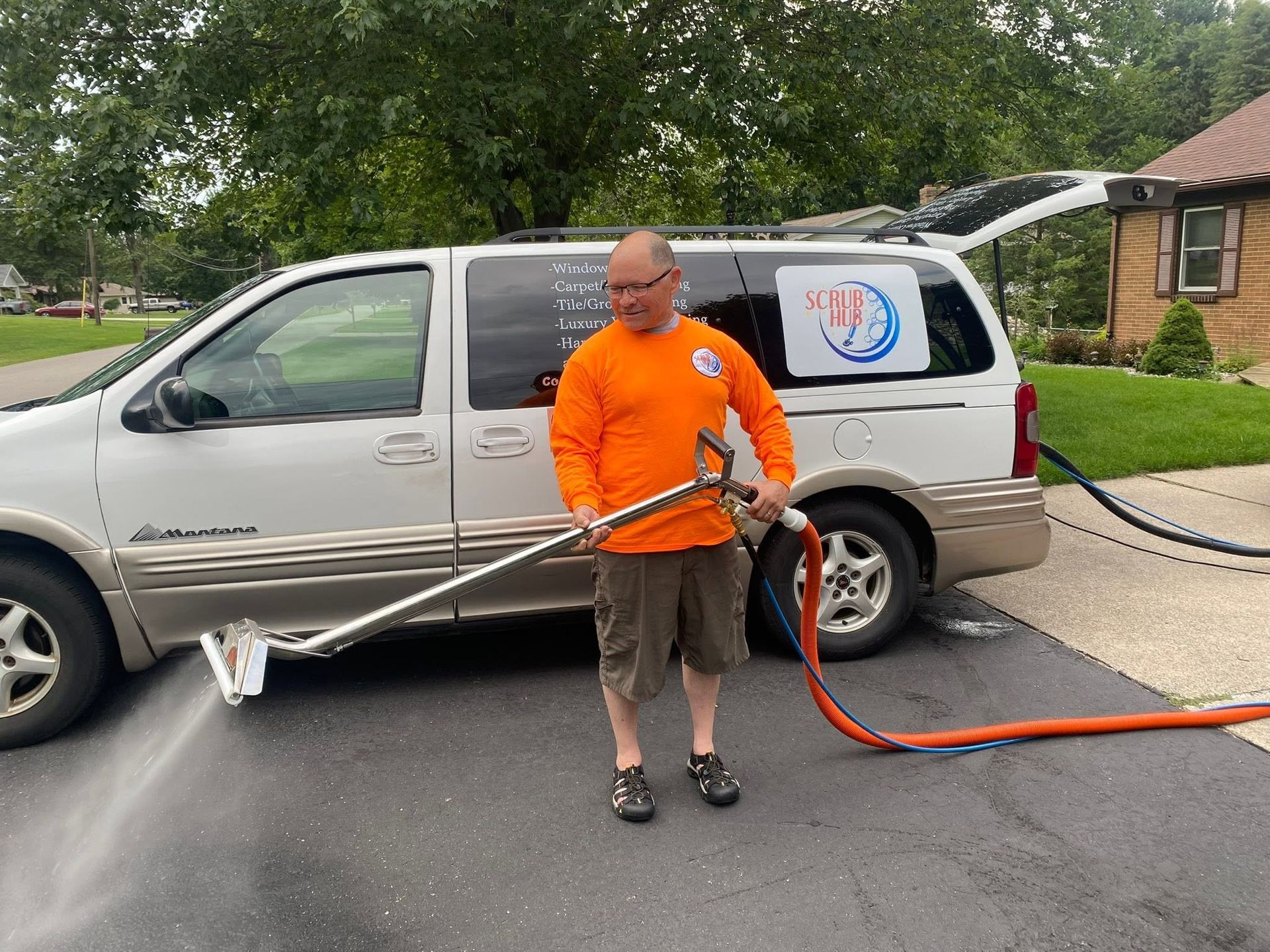 Man spraying driveways with a hose beside a silver minivan and orange hose on a residential street