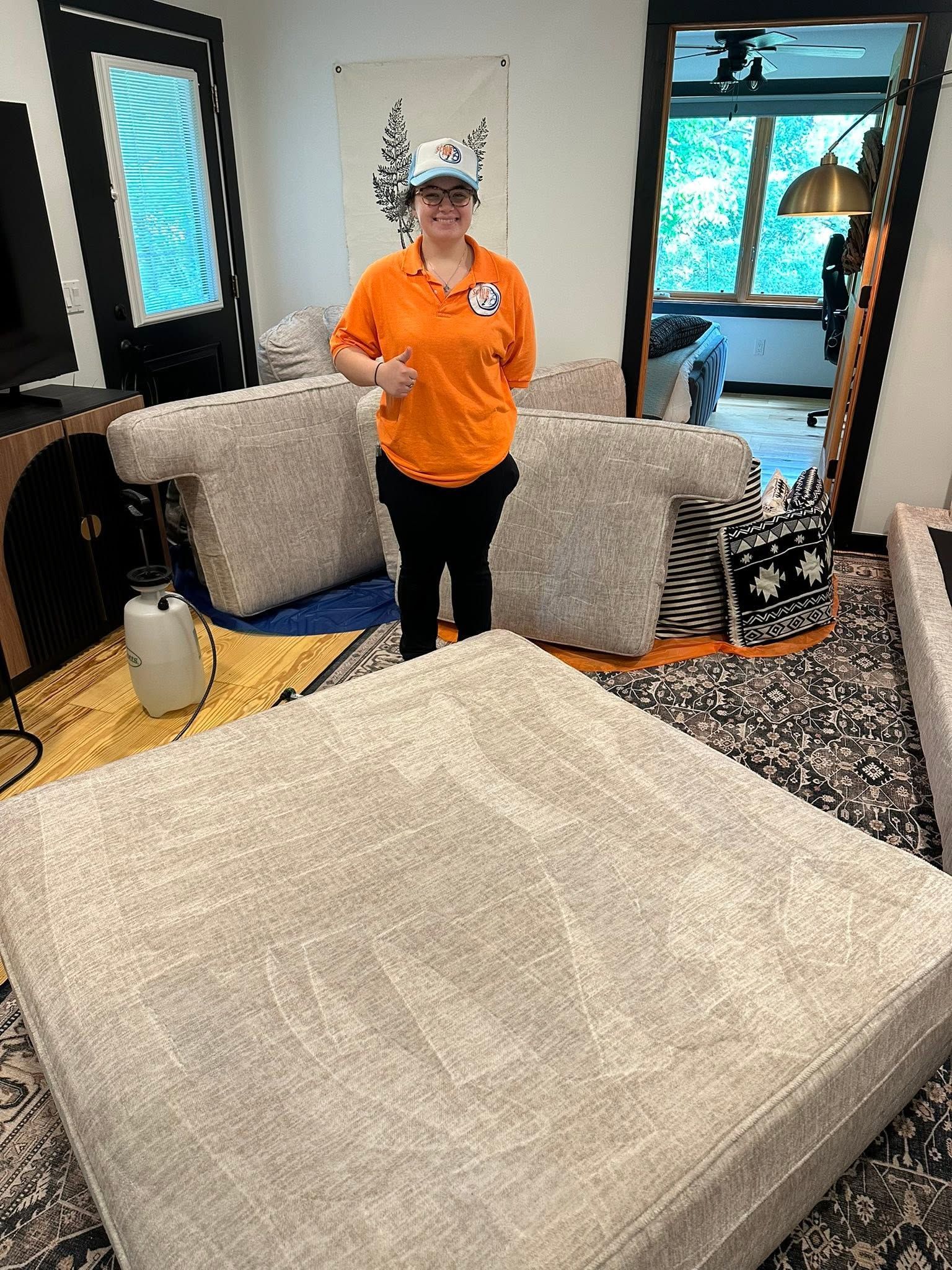 Person in orange shirt standing in a living room with gray sofa, patterned rug, and blue windows.