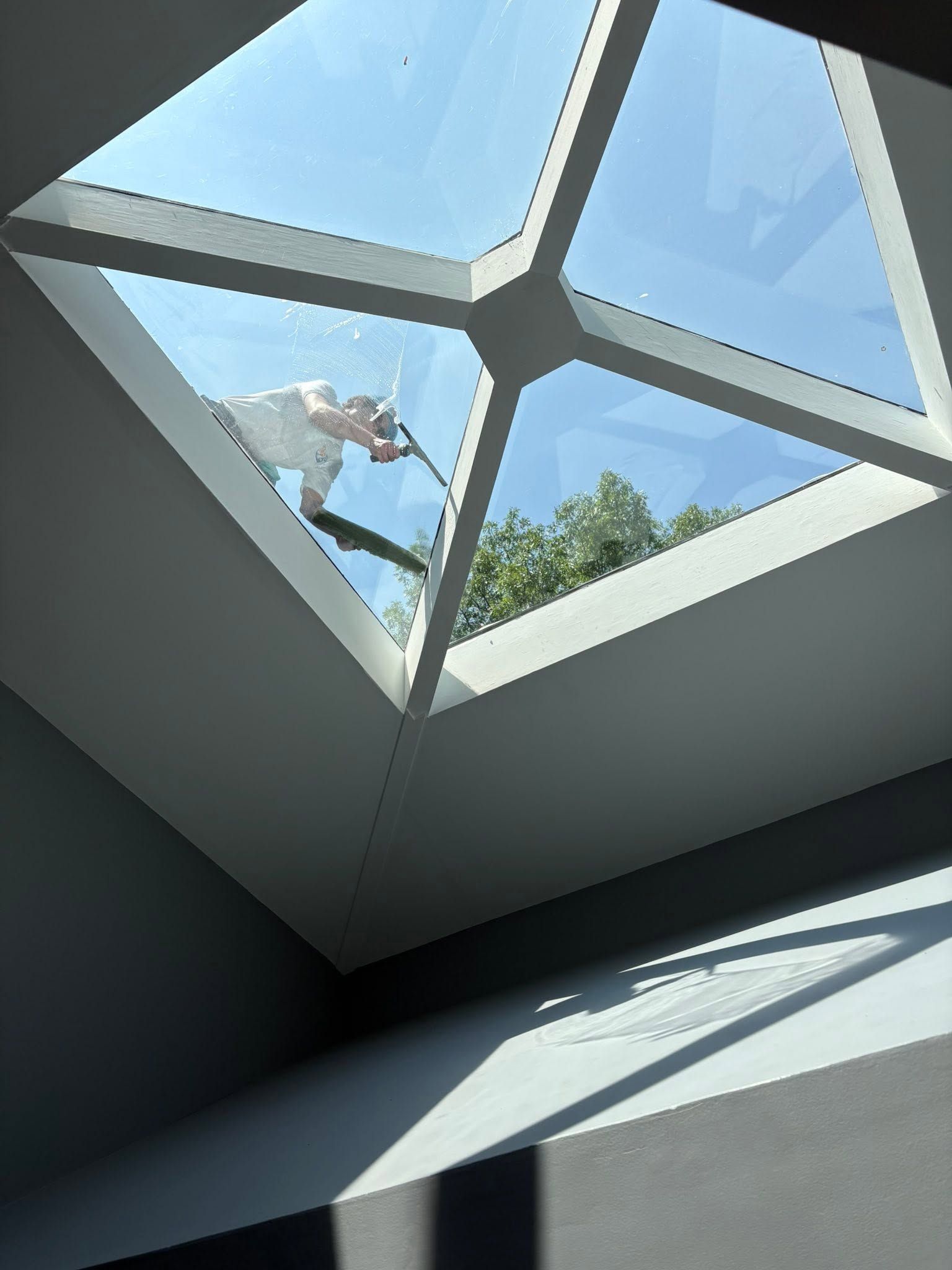 Person hanging from a rope near a modern skylight, with blue sky and trees visible above.