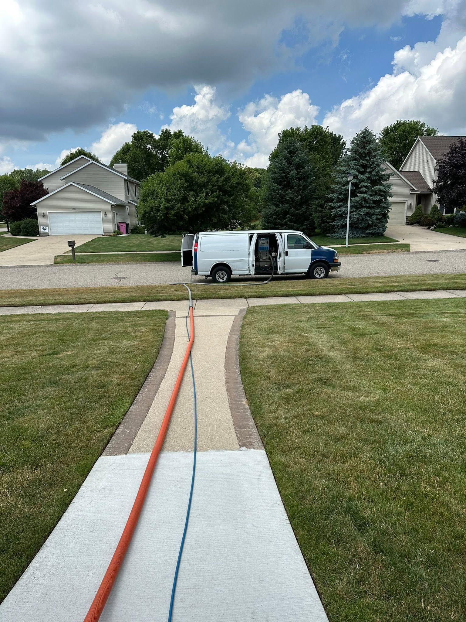 White work van parked across a suburban street, viewed from a front walkway under a partly cloudy sky.