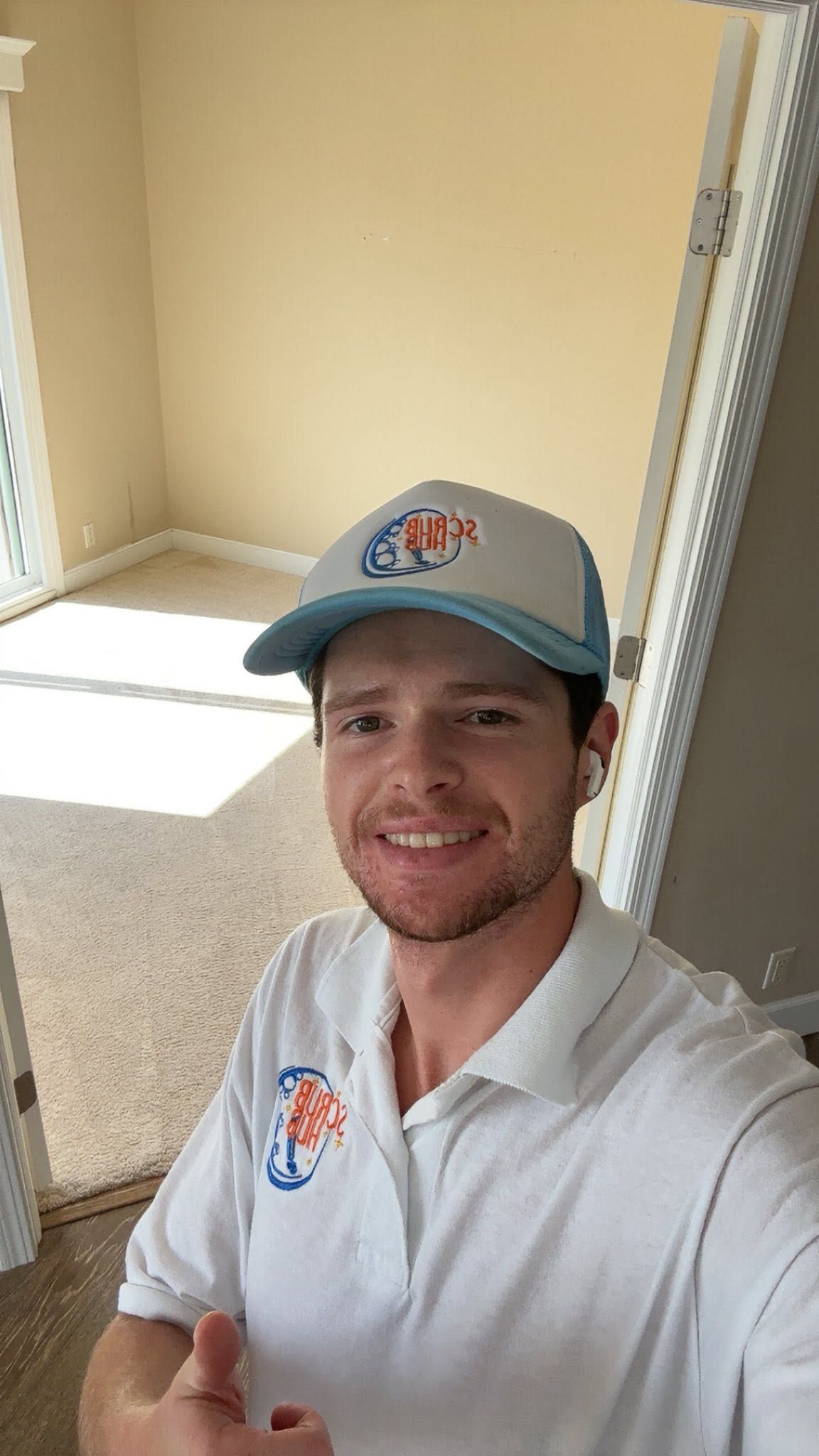 Person smiling in a white polo and gray cap, taking a selfie in a sunlit room with beige carpet.