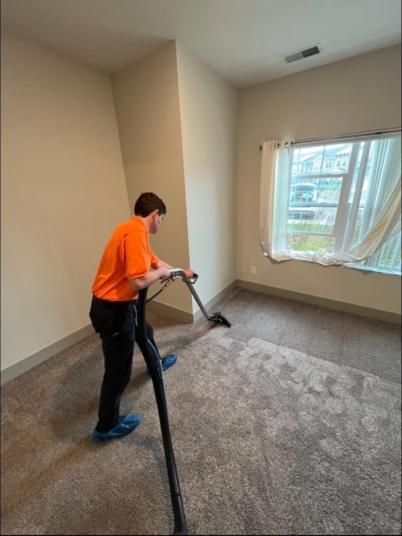 Person vacuuming a beige carpet in an empty room beside a sunlit window.