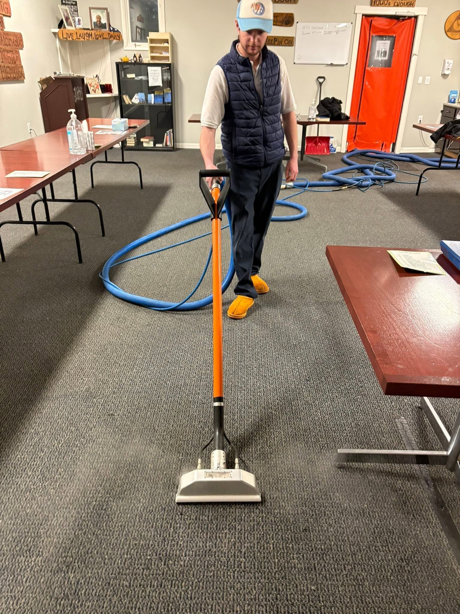 Person vacuuming carpet in a room with tables, chairs, and coiled blue hose nearby.