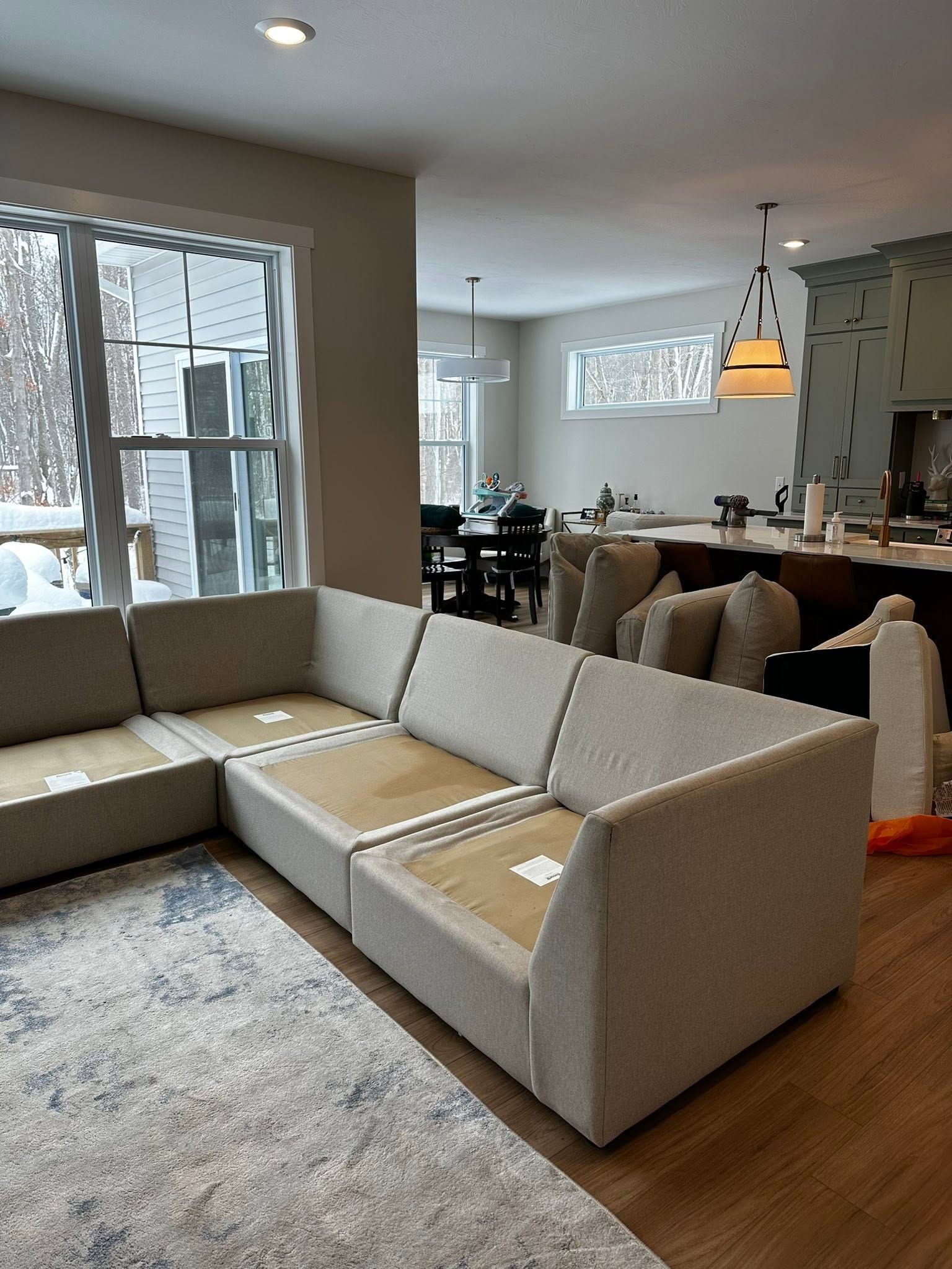 Modern living room with a beige sectional sofa, large windows, and a dining area in the background