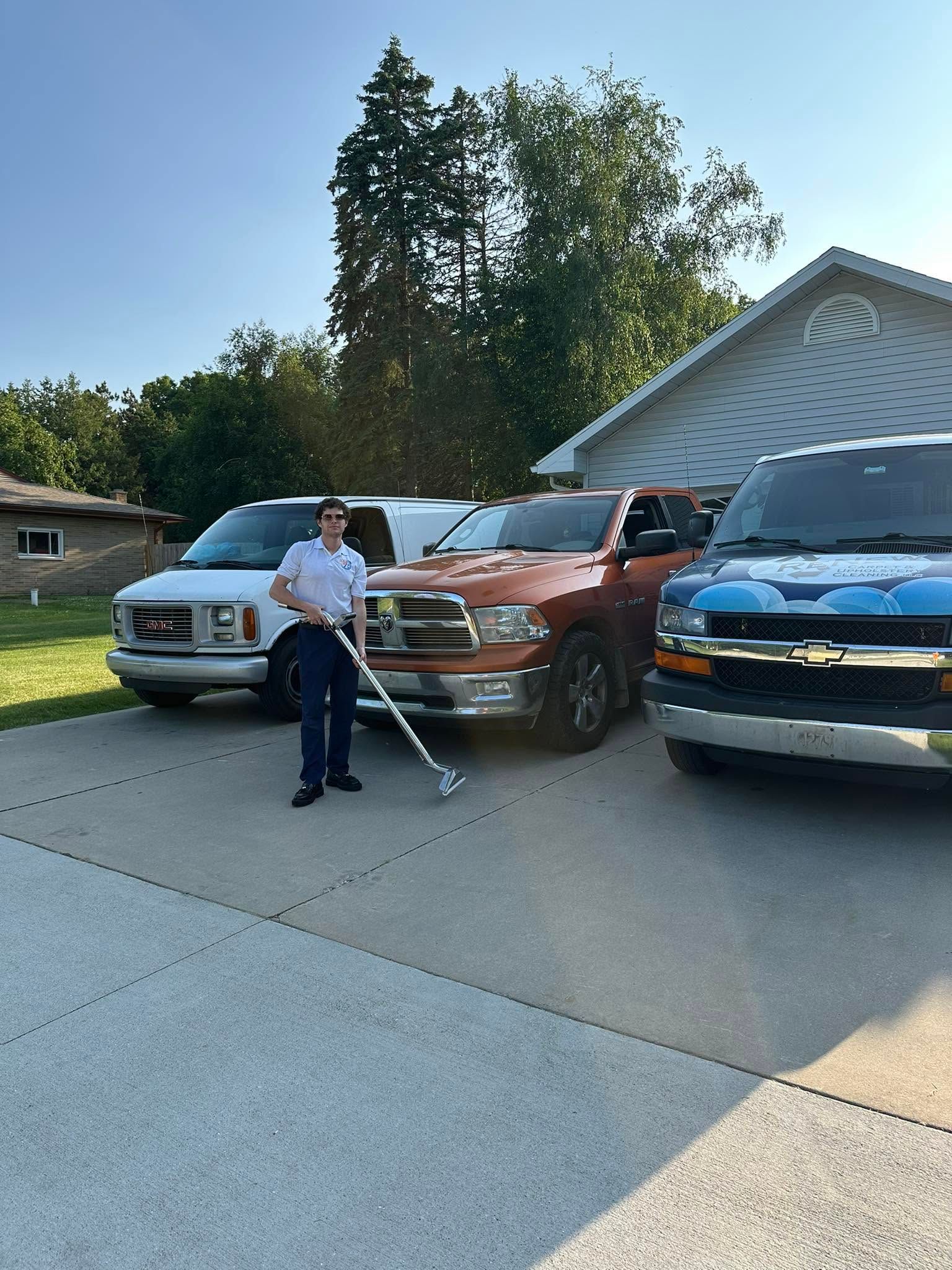 Person standing with a white cane in a driveway beside parked trucks and cars on a sunny day