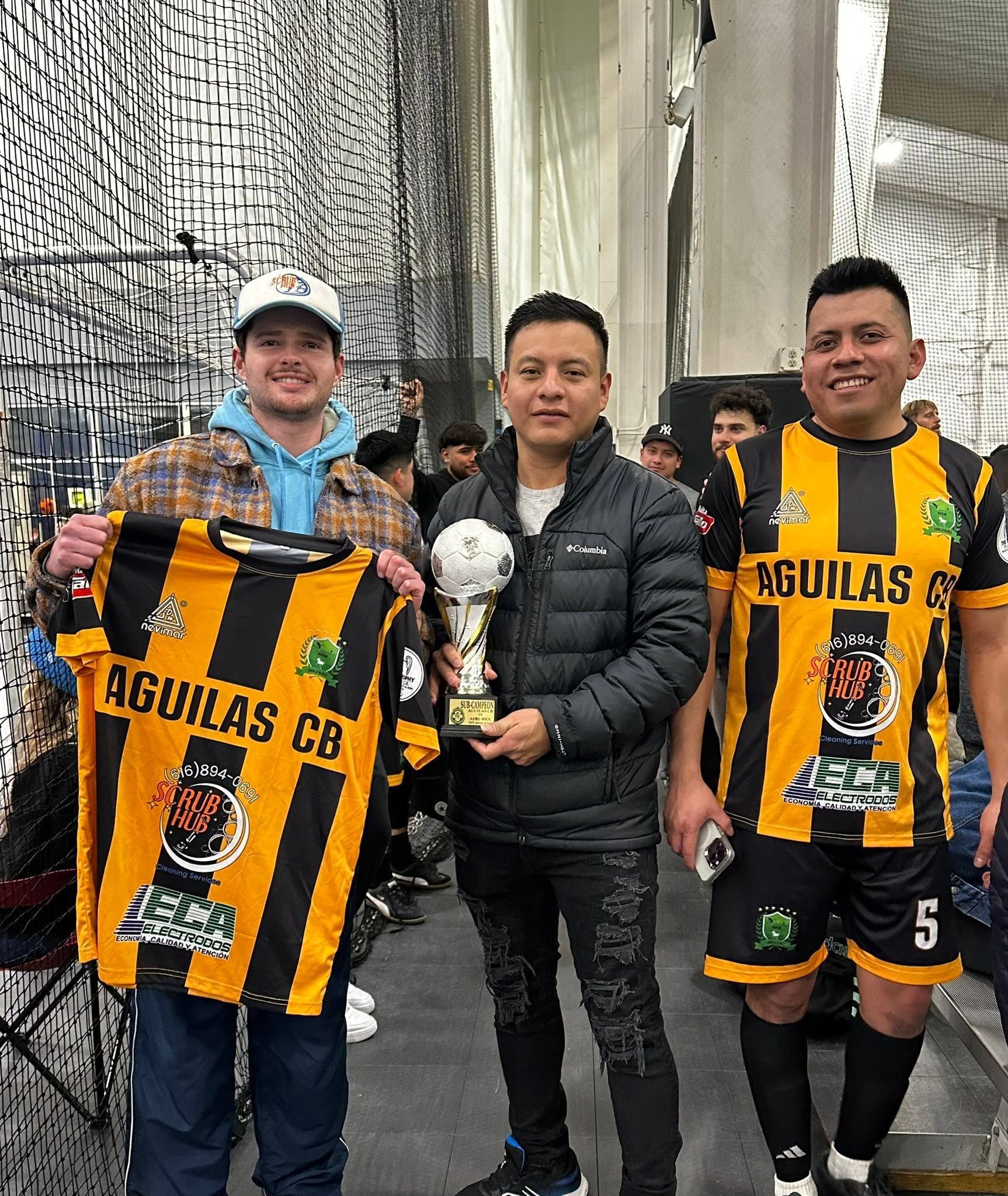 Three soccer fans pose holding striped Águilas FC jerseys in front of a netted wall.