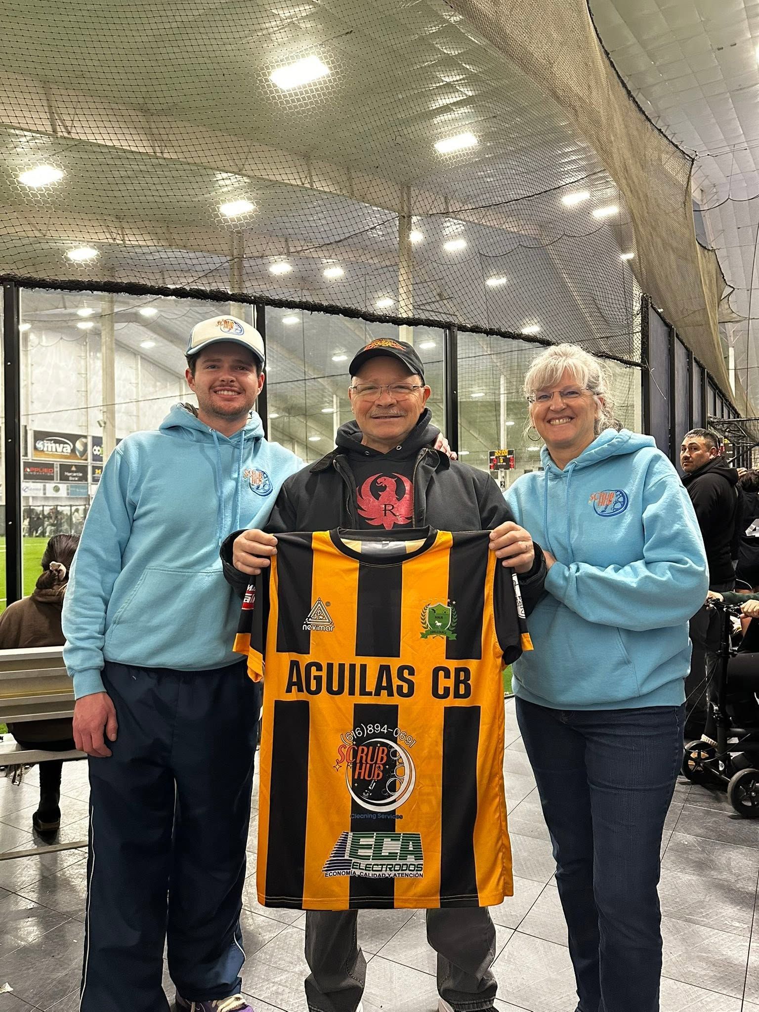 Three people holding a yellow-and-black Aguilas CB soccer jersey indoors.